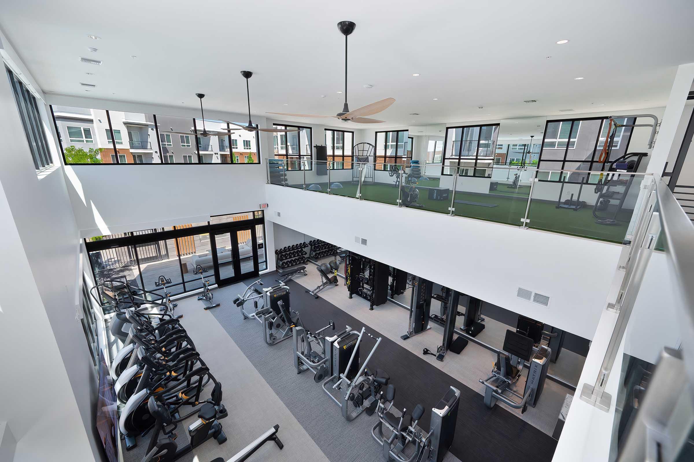 A spacious modern gym featuring multiple workout areas, including cardio machines, weightlifting equipment, and an open design with large windows. The two-level setup includes a second-floor balcony overlooking the main workout space, enhancing the overall bright and inviting atmosphere.
