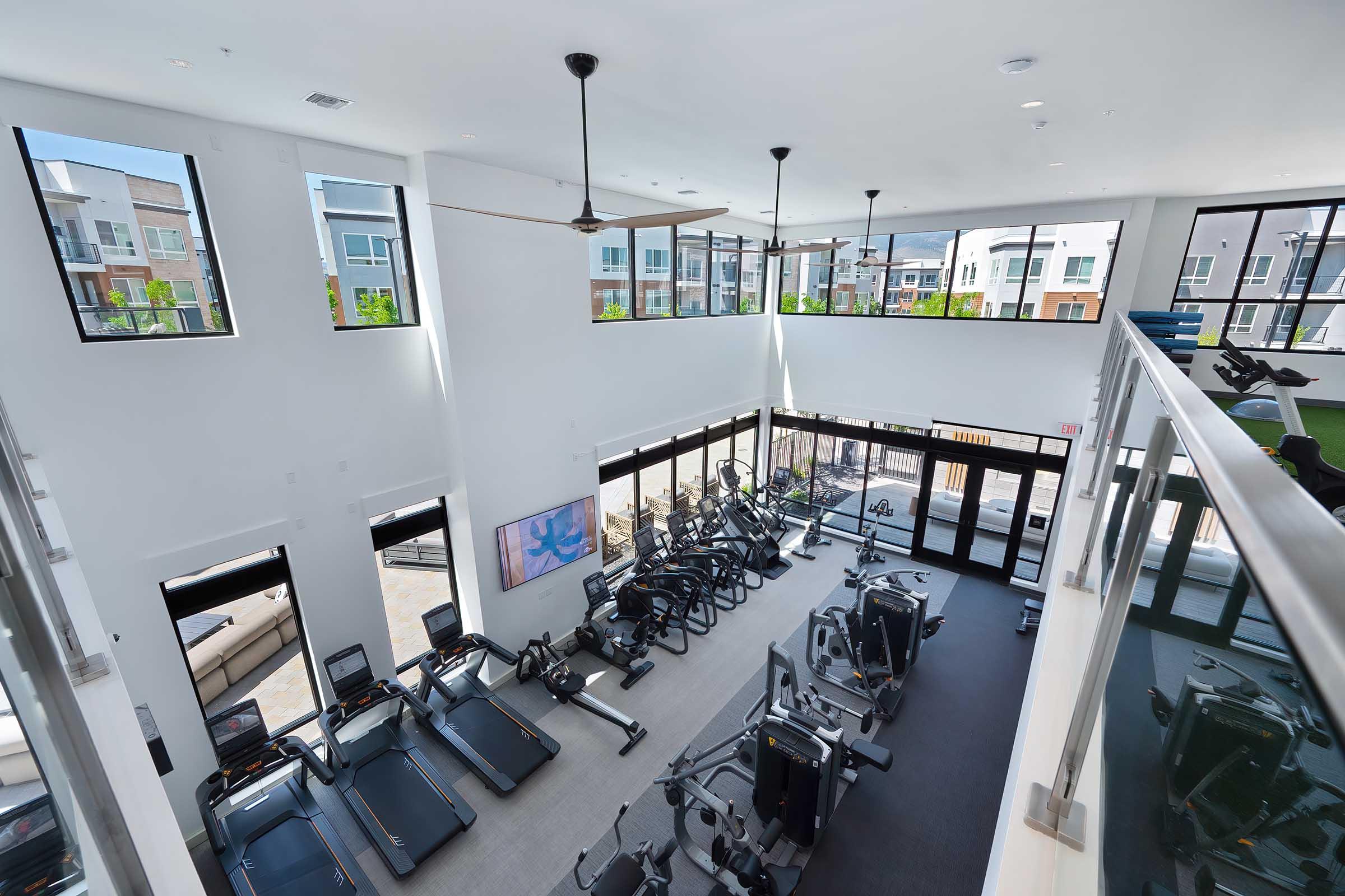 A bright and spacious fitness center featuring a variety of workout equipment, including treadmills and exercise machines. Large windows let in natural light, and there are modern ceiling fans. The design is open and contemporary, with a view of the outdoor area and additional workout spaces.