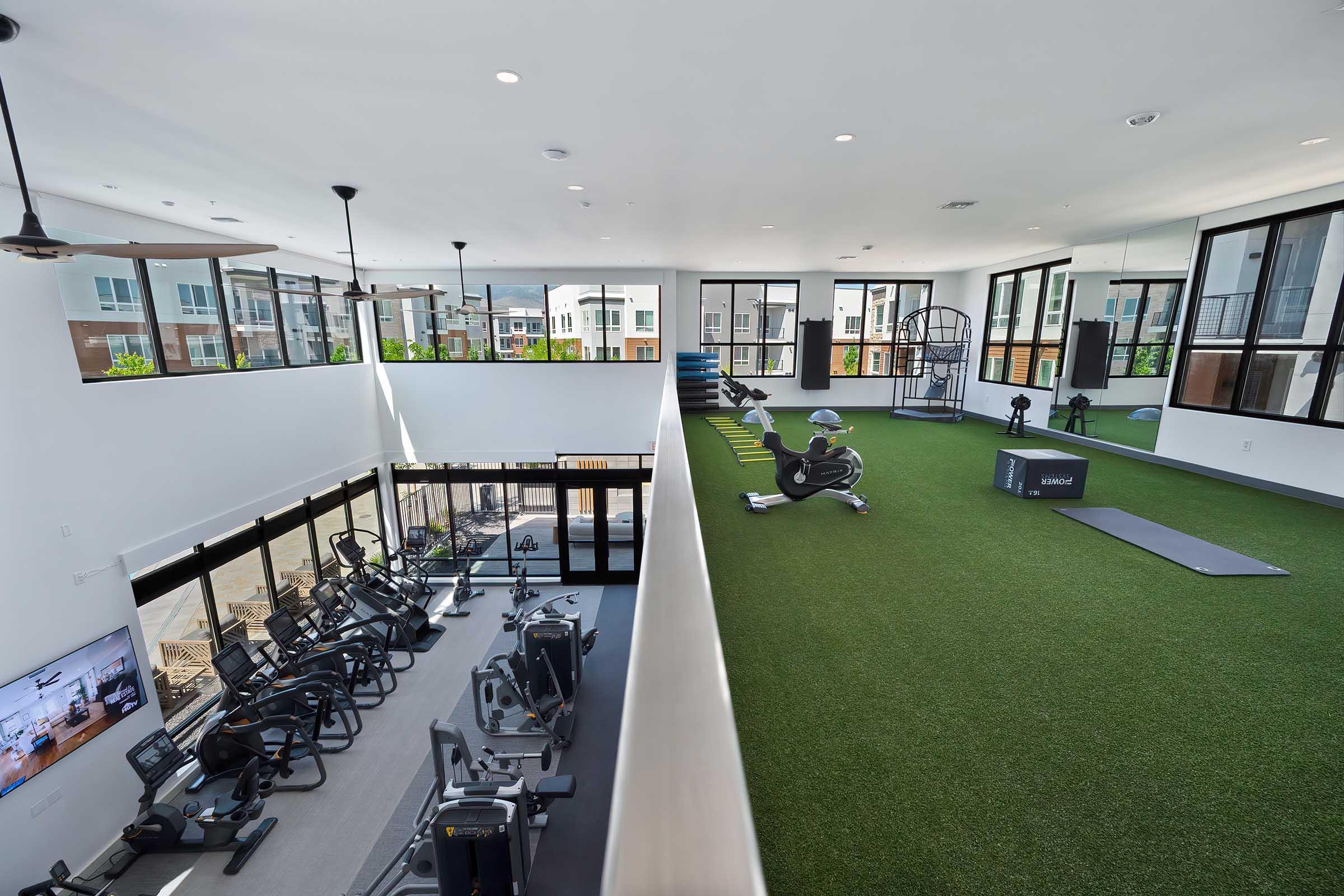 A modern fitness facility featuring a spacious layout with cardio equipment on one side and a training area with artificial turf on the other. Large windows allow natural light, and there's a mirrored wall for exercise reflection. Various workout equipment is available, creating an inviting workout environment.