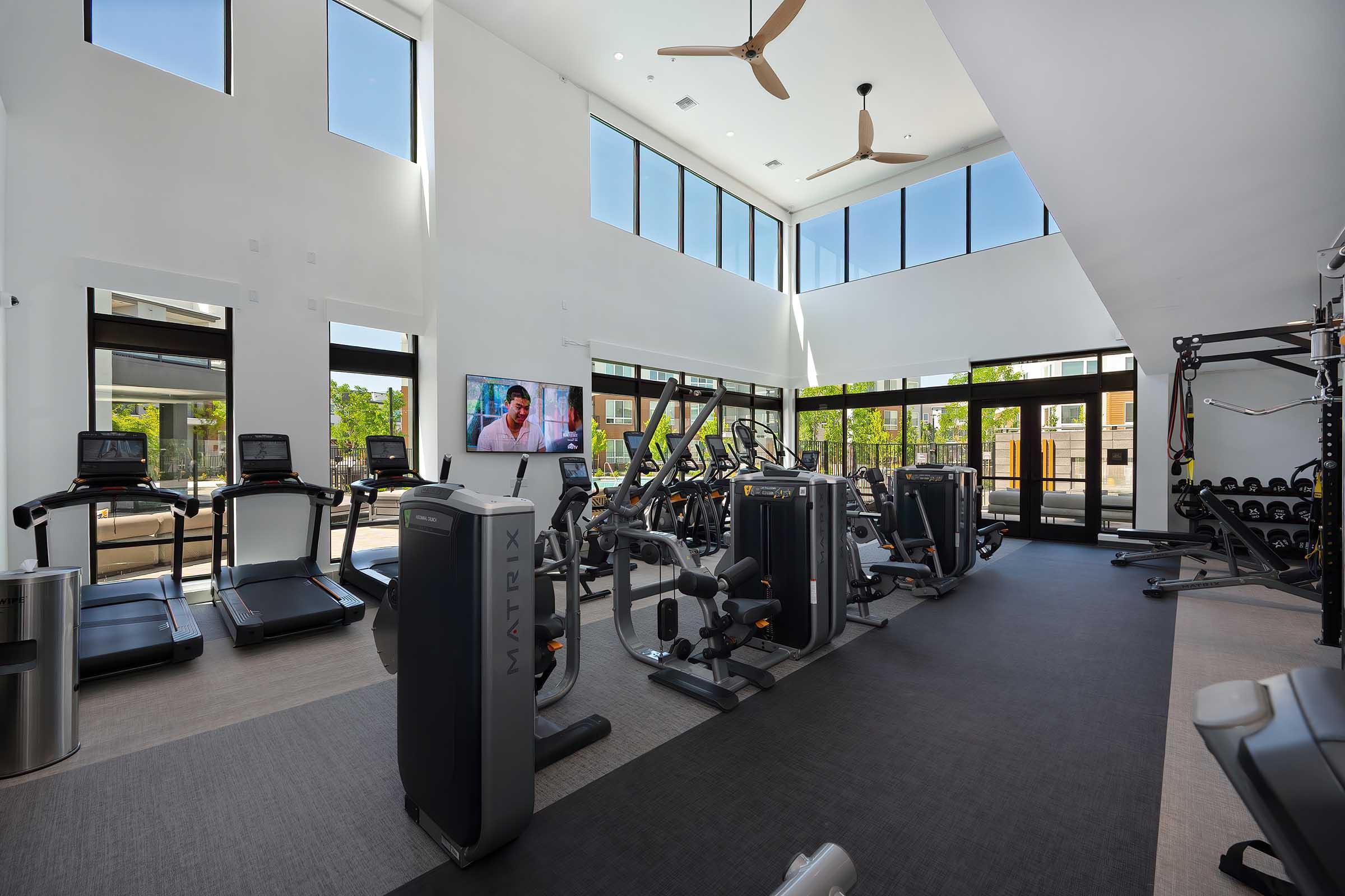 A spacious, modern gym featuring several cardio machines, including treadmills and stationary bikes. Large windows allow natural light to fill the room, and a wall-mounted TV plays a fitness program. The area includes weight training equipment and a sleek, contemporary design.