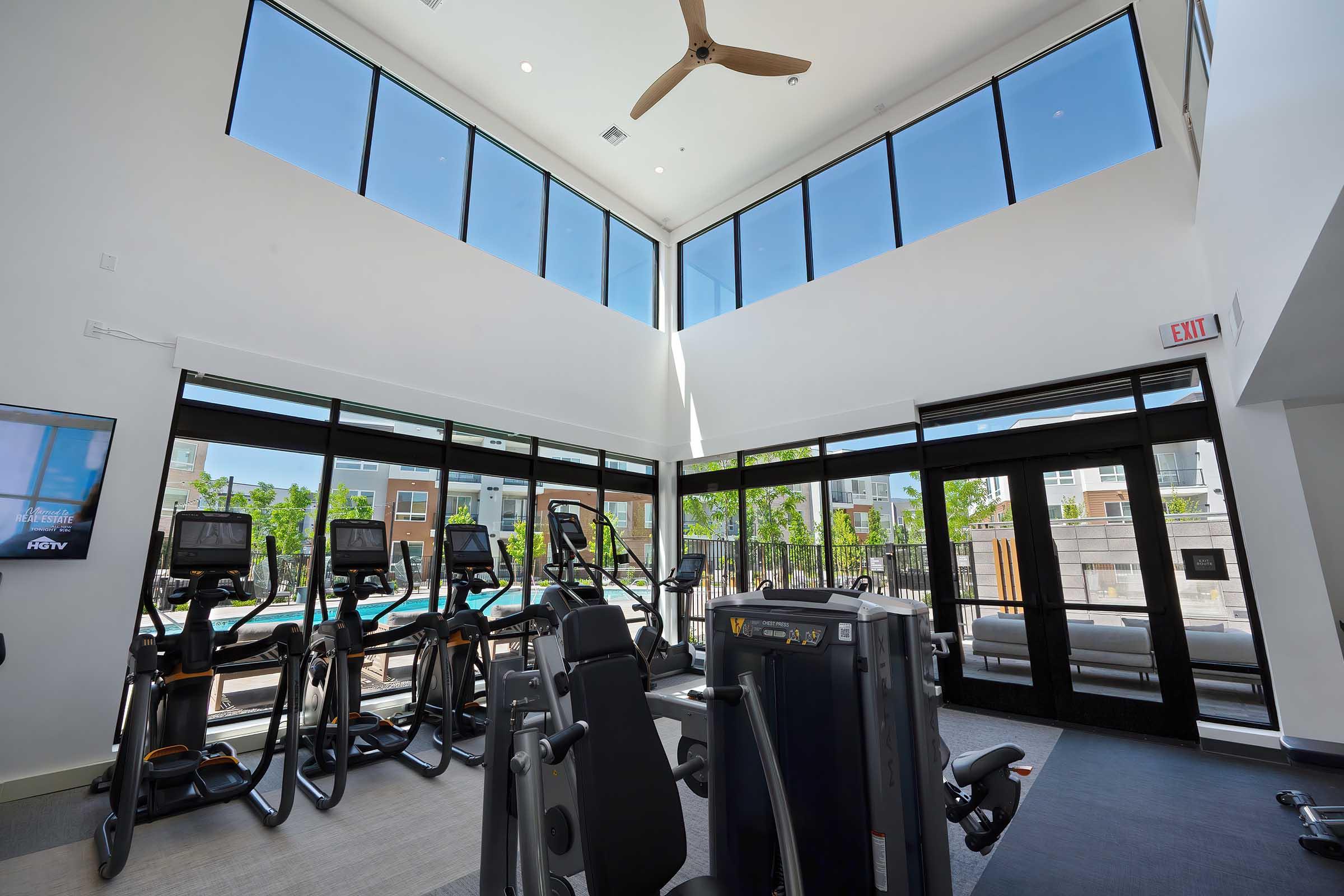 Modern gym interior with large windows allowing natural light. Equipped with elliptical machines and weight machines. There's a ceiling fan and a view of a swimming pool area outside. The space is bright and airy, designed for fitness and wellness.