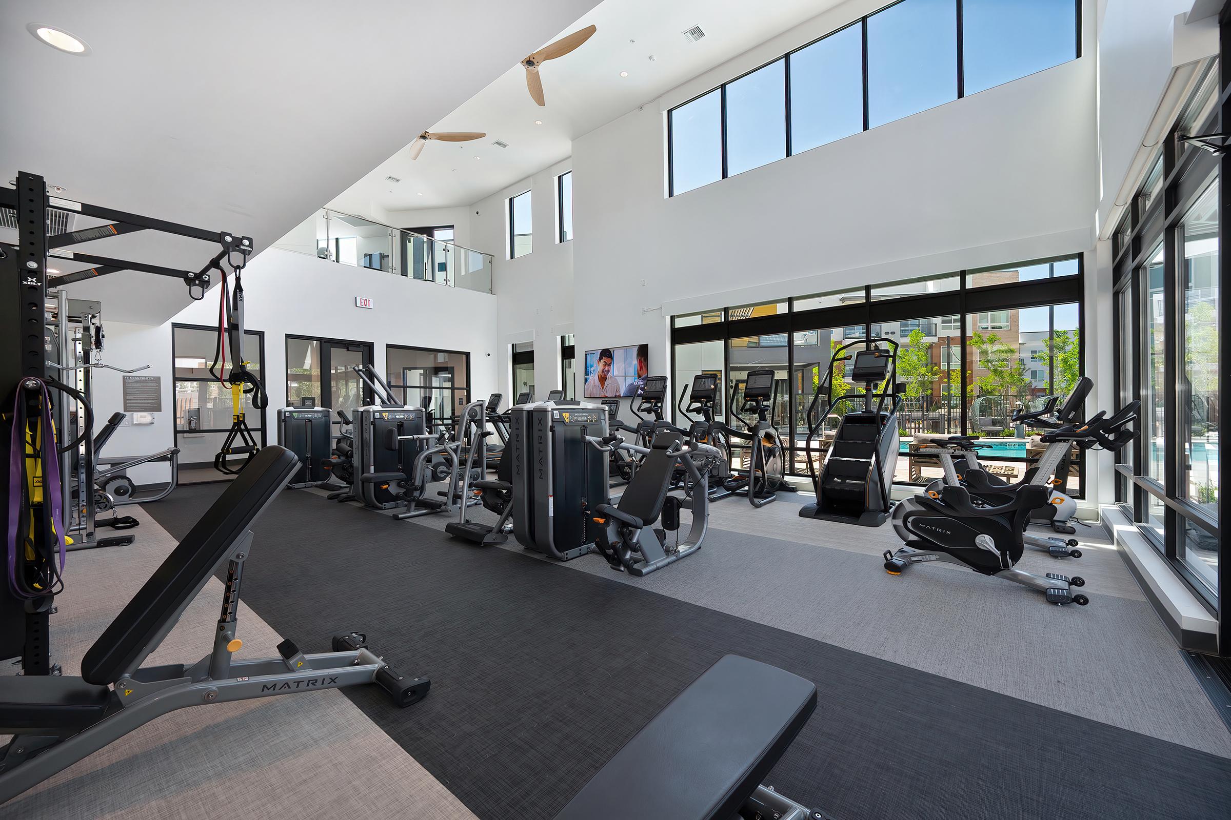 A modern gym interior with large windows, featuring various exercise equipment like treadmills, stationary bikes, and weight machines. Natural light fills the space, creating a bright and inviting atmosphere for workouts. A television screen is mounted on the wall, adding to the gym's functionality.