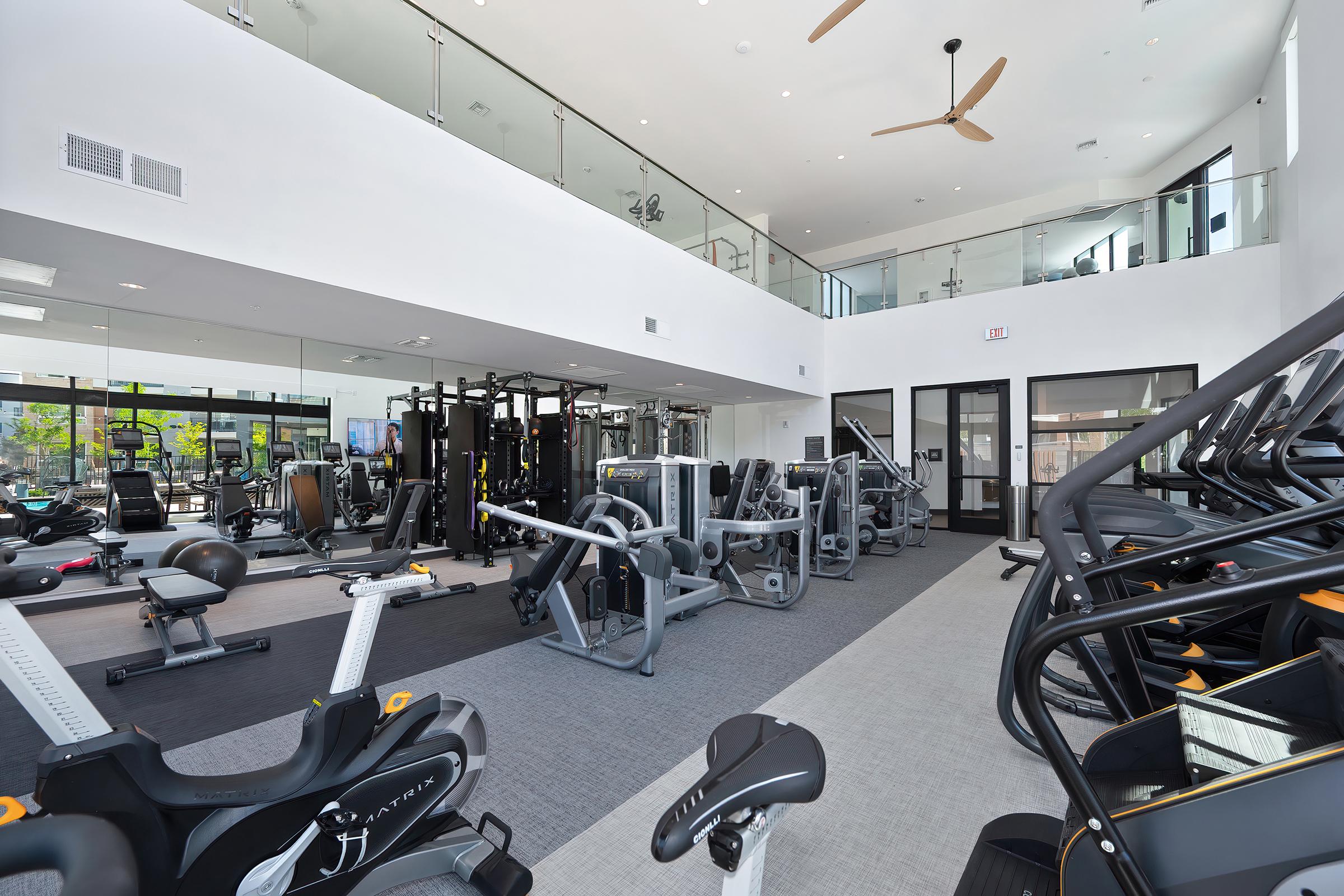 Modern gym interior featuring various workout machines and equipment. The space is well-lit with large windows and high ceilings, showcasing a variety of exercise machines, including treadmills and weightlifting stations. A sleek, contemporary design enhances the active atmosphere.