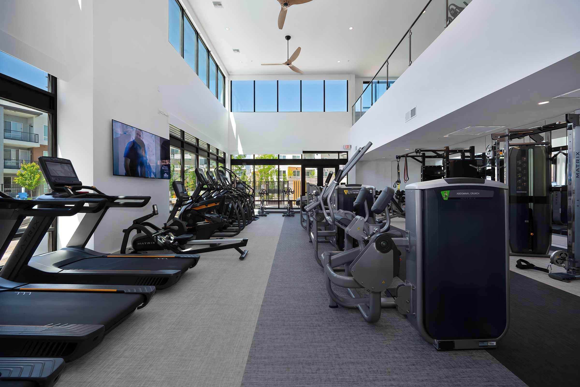 A bright and modern gym interior featuring various exercise equipment. Treadmills and stationary bikes are lined up along one side, while weight machines and strength training equipment are positioned along the opposite side. Large windows provide natural light, creating an inviting atmosphere.