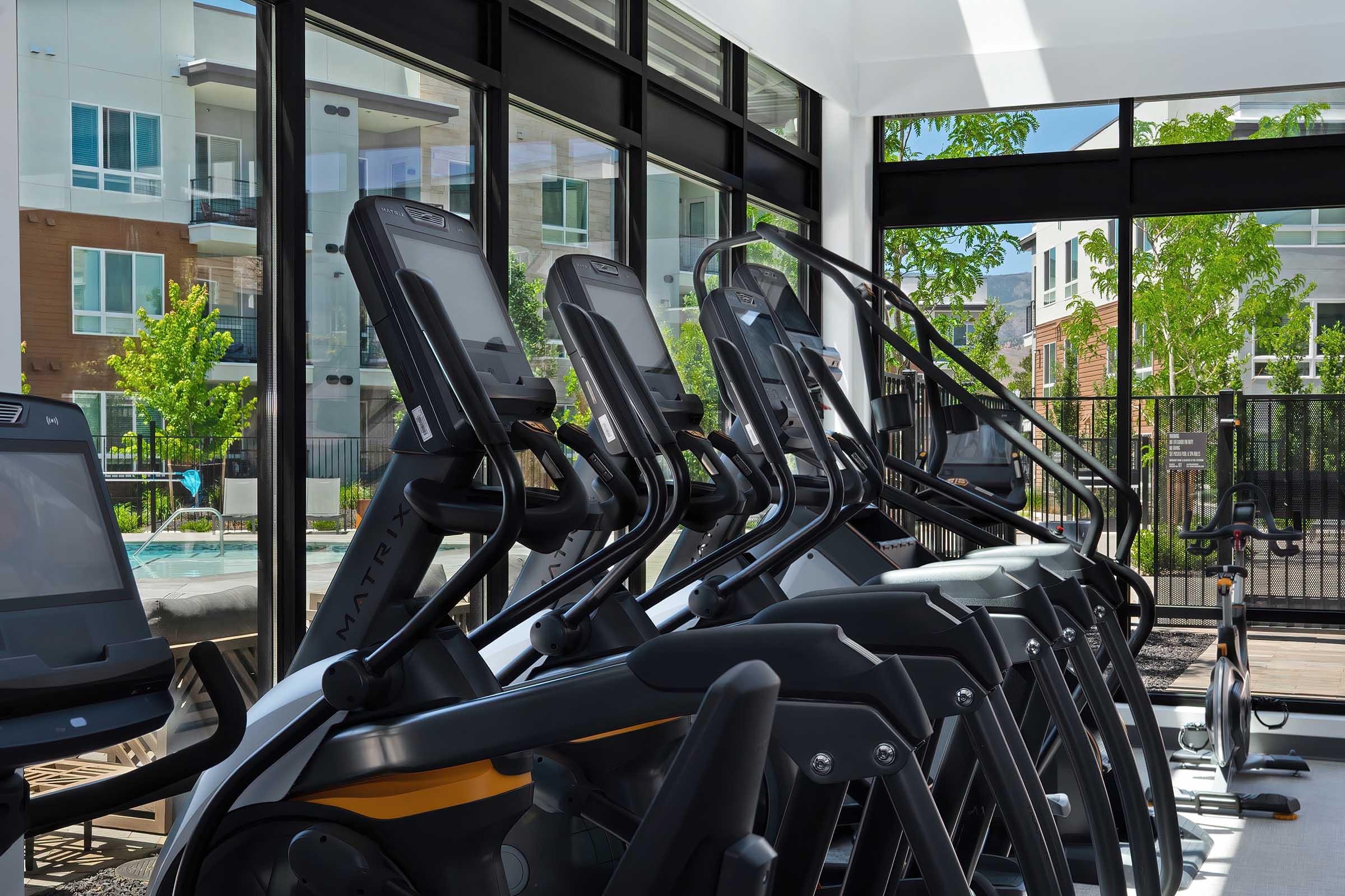 A modern fitness center featuring multiple elliptical machines lined up near large windows, allowing natural light to illuminate the space. The view outside includes green trees and an outdoor area, creating a refreshing atmosphere for workouts.