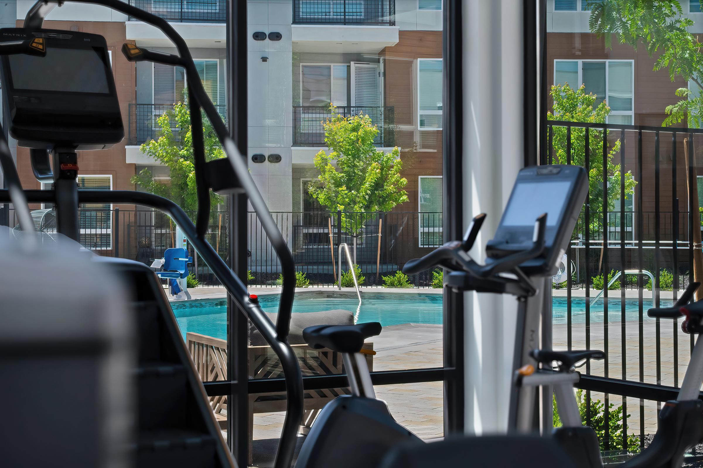 A bright fitness area with exercise equipment, like treadmills and bikes, facing large windows that overlook a swimming pool and lush greenery outside. The pool area features deck chairs and a fenced perimeter, creating a refreshing and inviting atmosphere for exercise and relaxation.