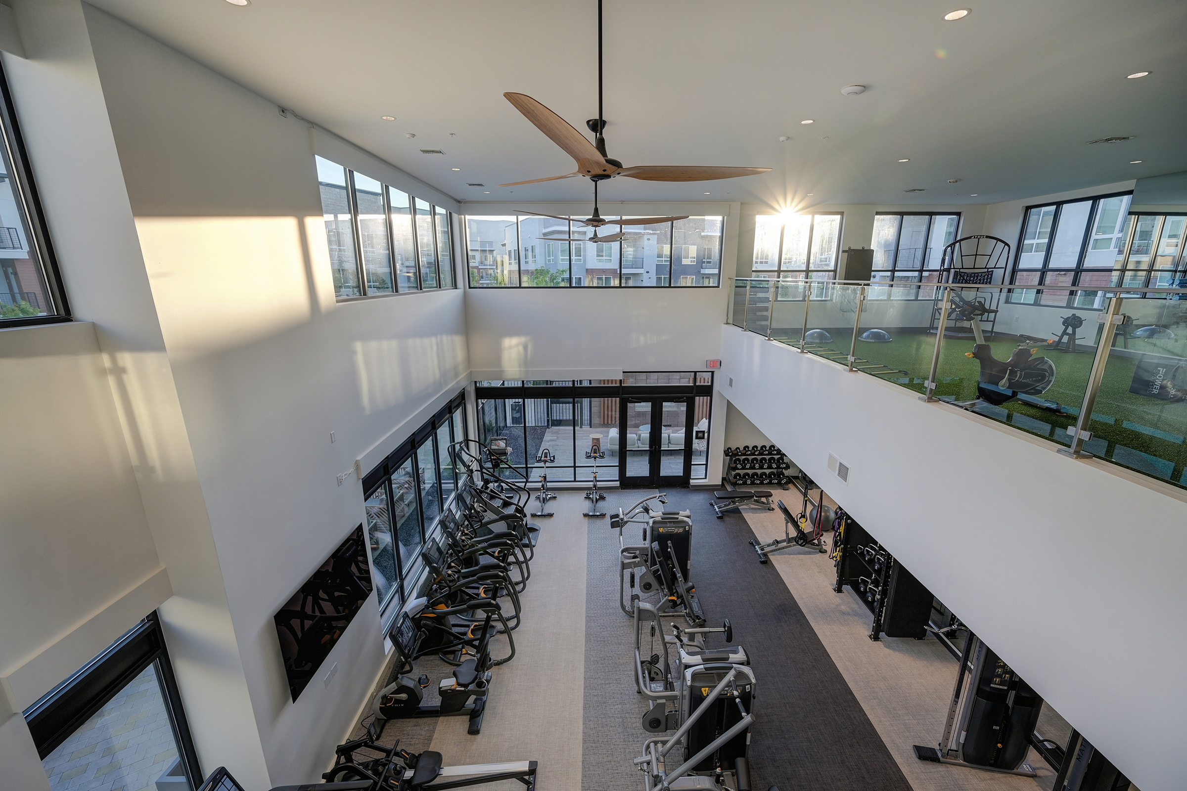 Bright and modern gym interior featuring cardio machines and weightlifting equipment. Large windows allow natural light to fill the space, with a view of outdoor fitness areas. A ceiling fan is visible, and the design emphasizes an open, spacious feel.