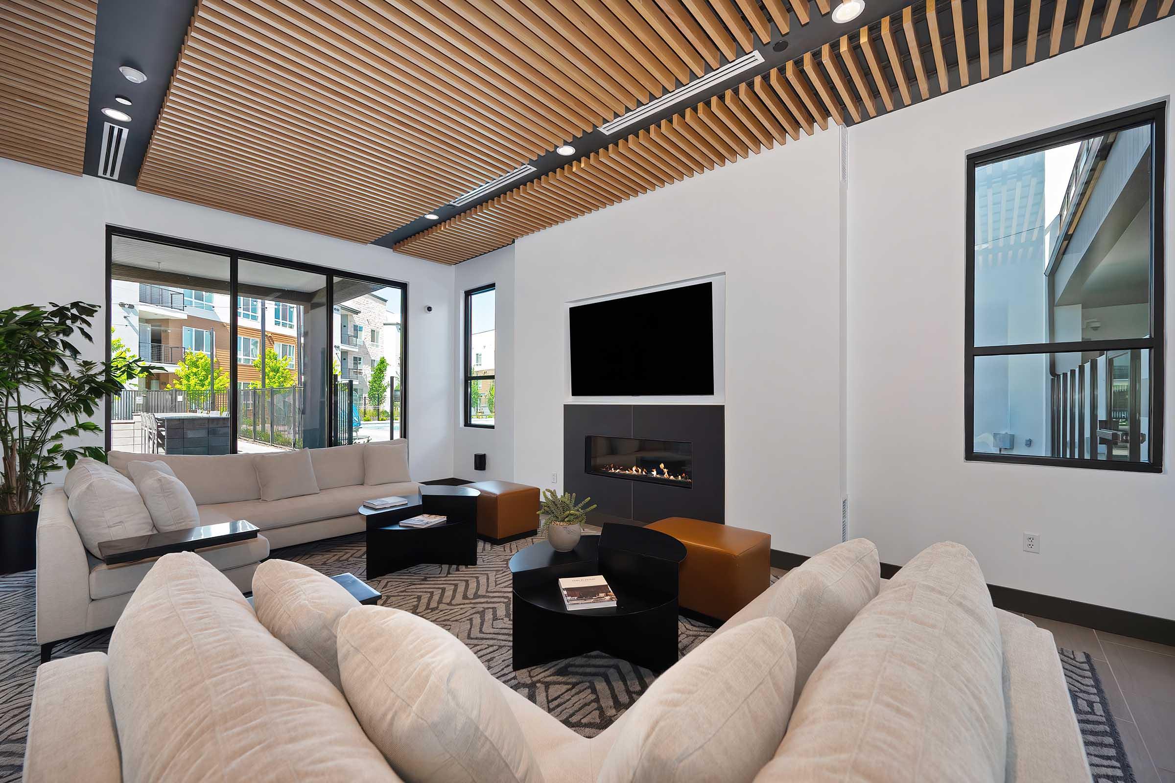 Modern living room featuring a cozy seating area with a large beige sectional sofa, a coffee table, and a sleek black media console with a TV. The room has large windows providing natural light, wooden ceiling details, and a minimalist aesthetic with green plants and decorative accents.