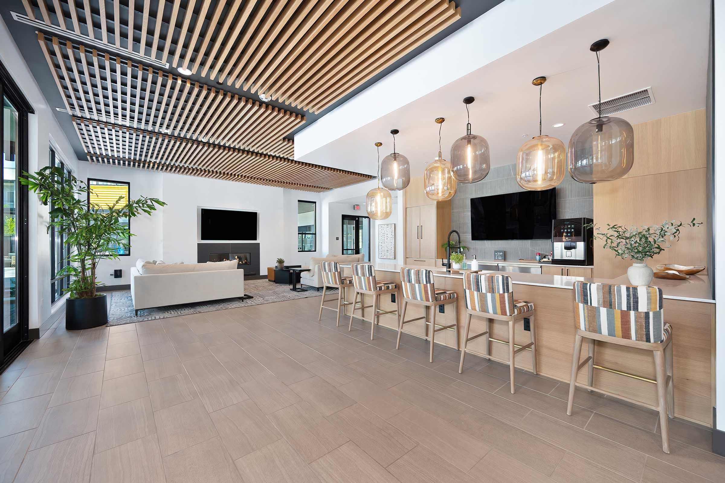 A modern, open-concept lounge and kitchen area featuring wooden ceiling panels, stylish pendant lighting, a large flat-screen TV, and a cozy seating arrangement. The space includes a breakfast bar with high-backed chairs and a decorative plant, creating a welcoming atmosphere.