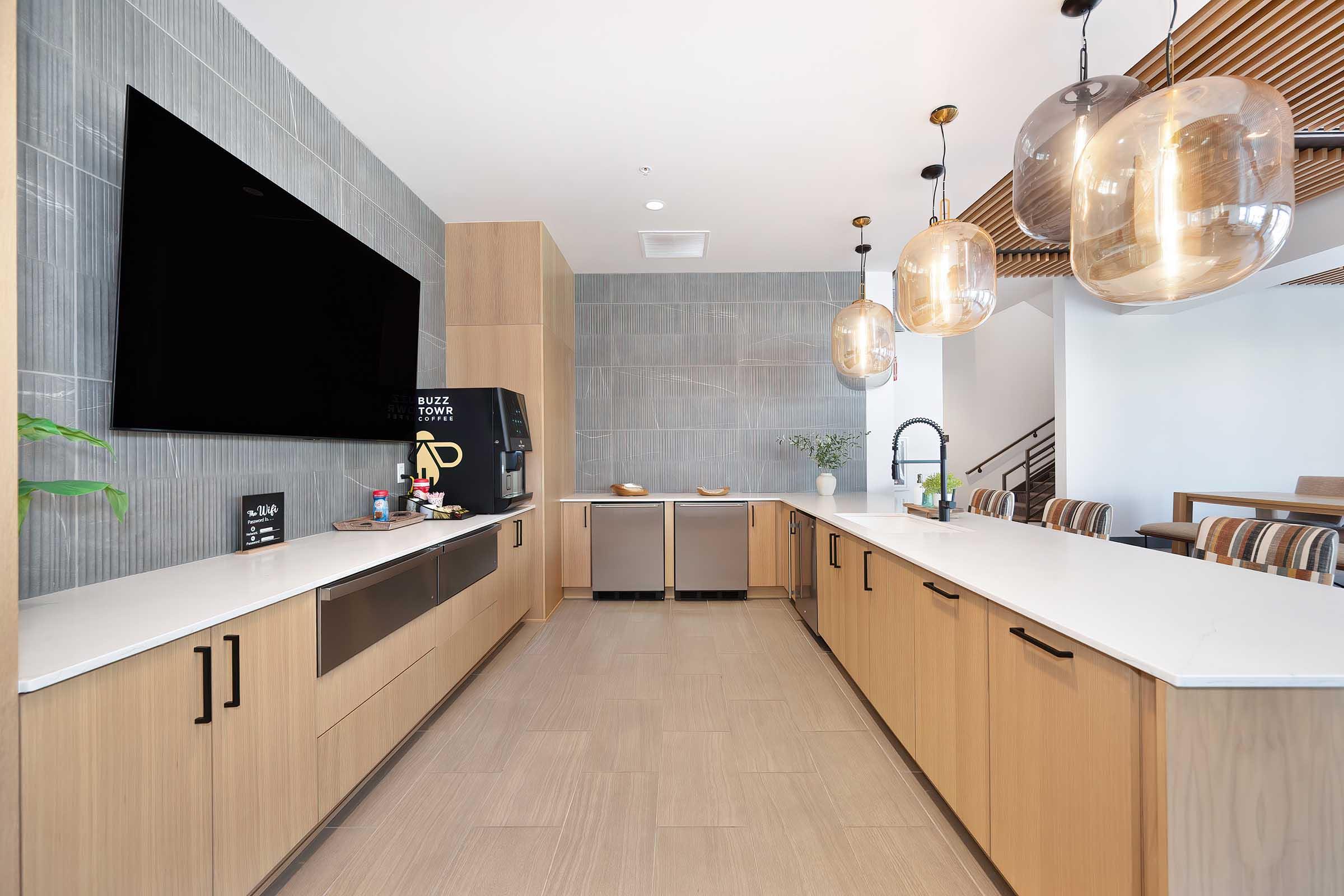 Modern kitchen featuring light wood cabinetry, a large flat-screen TV mounted on the wall, a coffee machine, and sleek stainless steel appliances. Pendant lights hang above a spacious countertop, with a neutral color palette and stylish seating in the background, creating a bright and inviting atmosphere.