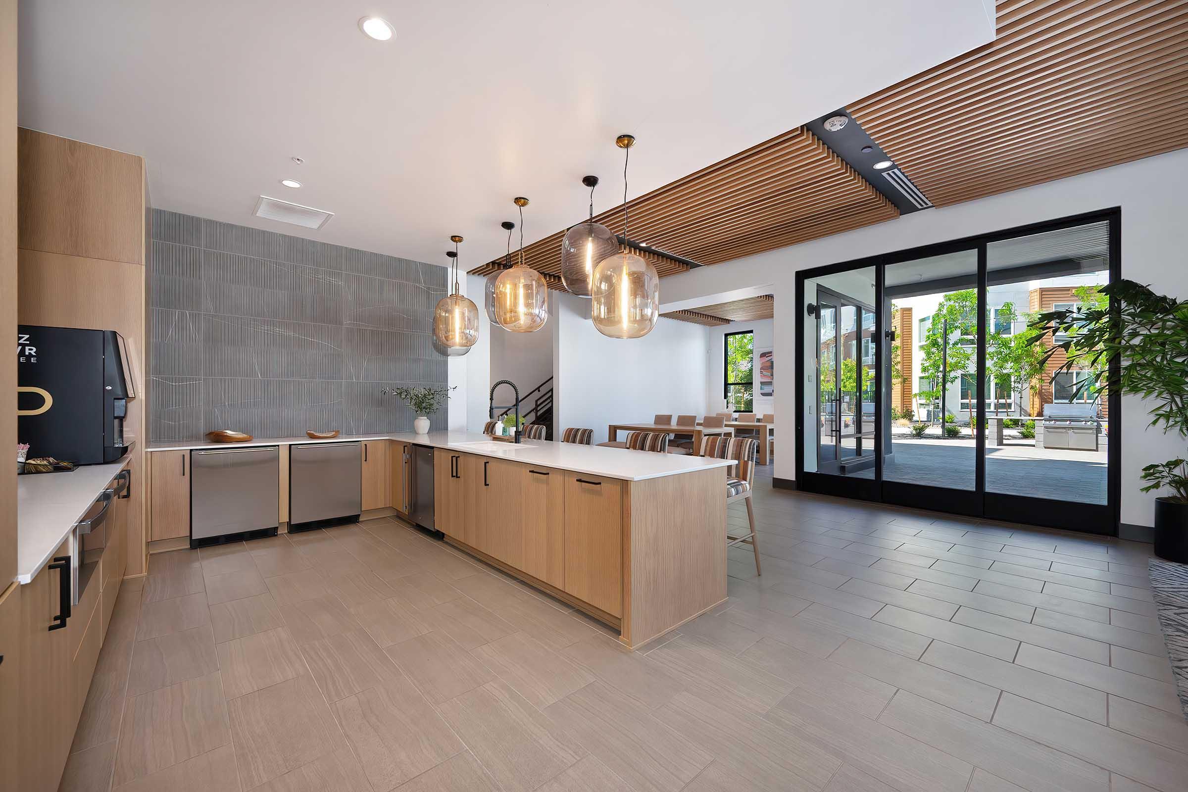 Modern kitchen with light wooden cabinetry, an island with a white countertop, and elegant pendant lighting. The space features large glass doors opening to an outdoor area, with a minimalist design and natural light streaming in. There are plants and dining areas visible, creating an inviting atmosphere.