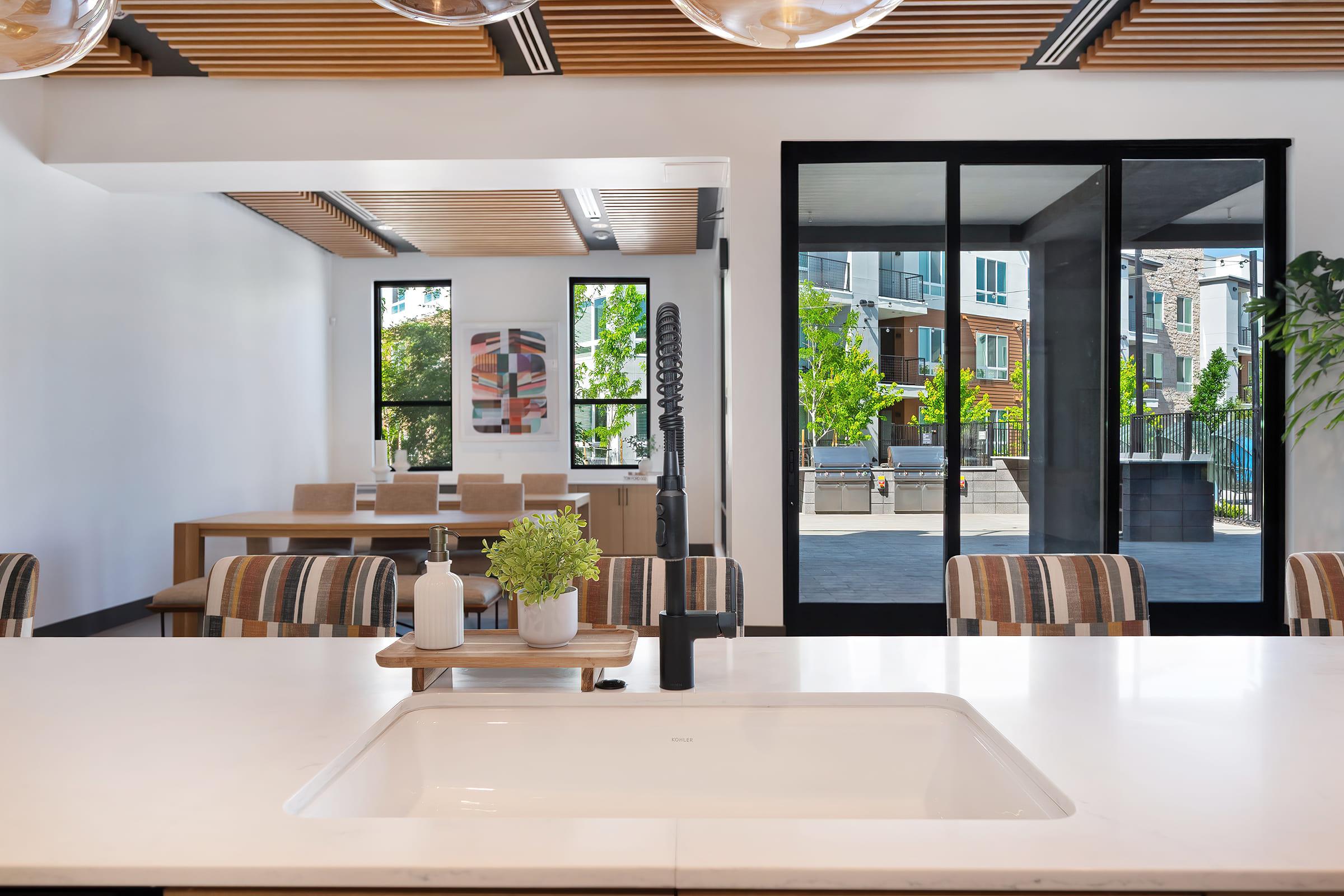 Modern kitchen with a large white countertop and a sleek black faucet. In the background, a dining area features a wooden table with striped chairs. Large windows allow natural light to flood in, showcasing views of an outdoor space with greenery. Contemporary design elements are prominent throughout the room.