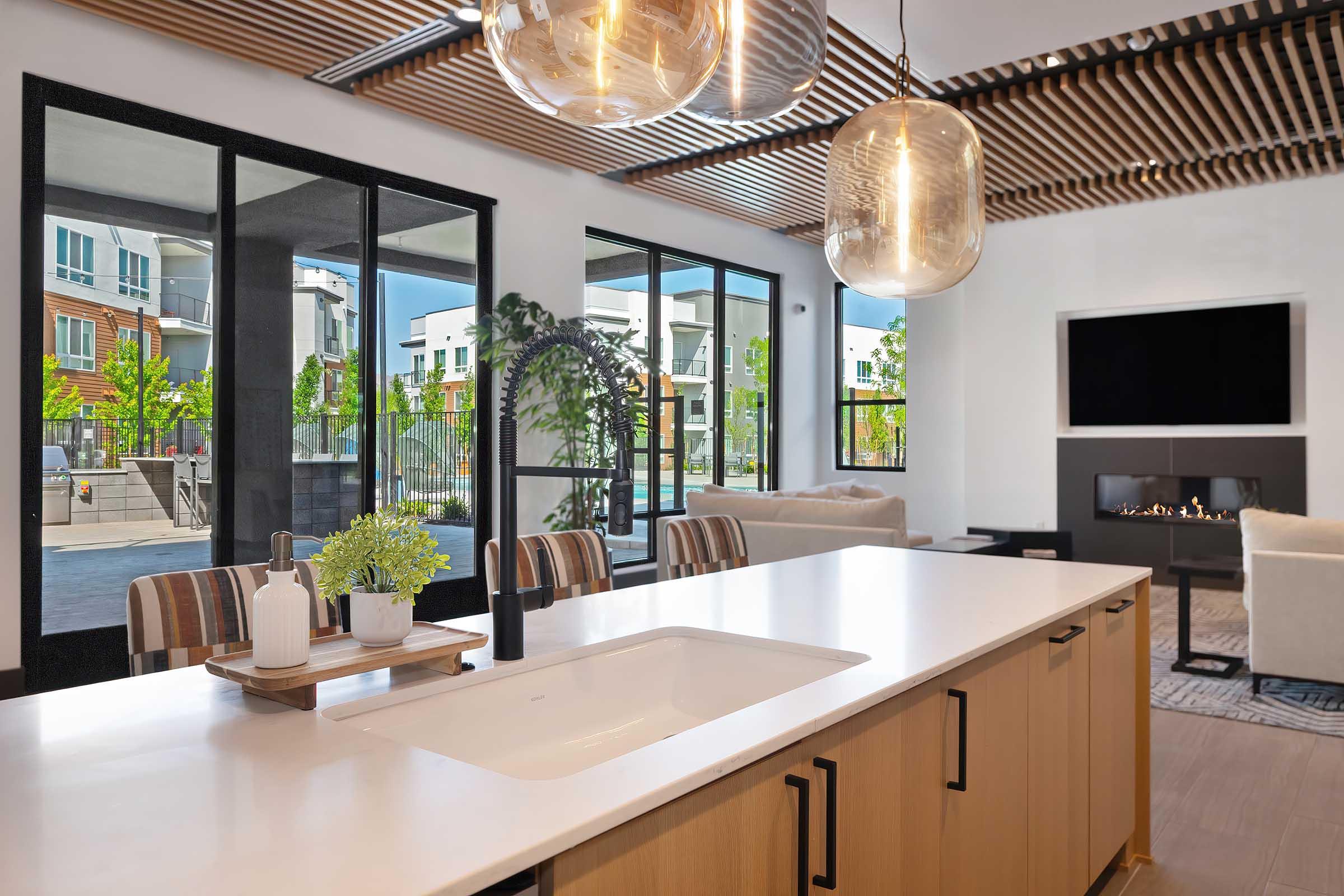 A modern interior space featuring a kitchen with a white countertop and wood cabinetry. Large windows allow natural light, showcasing a view of greenery outside. Stylish light fixtures hang from the ceiling, and there is a cozy seating area with a fireplace and a television on the wall.