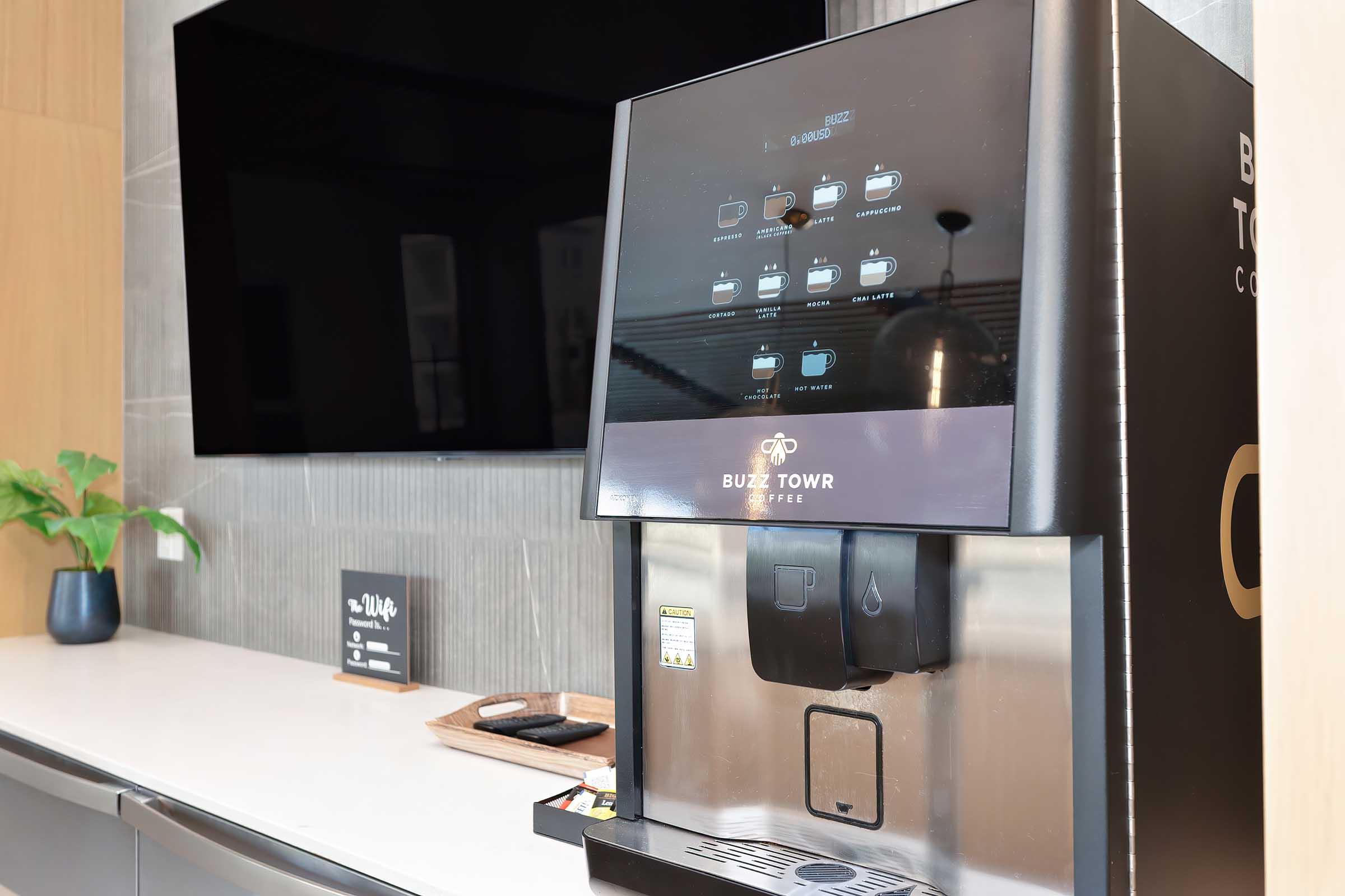 A coffee machine with a black and silver design, featuring a digital display panel with various drink options. It is situated on a countertop with a monitor in the background and a small potted plant nearby. A sign and some items are visible on the countertop.
