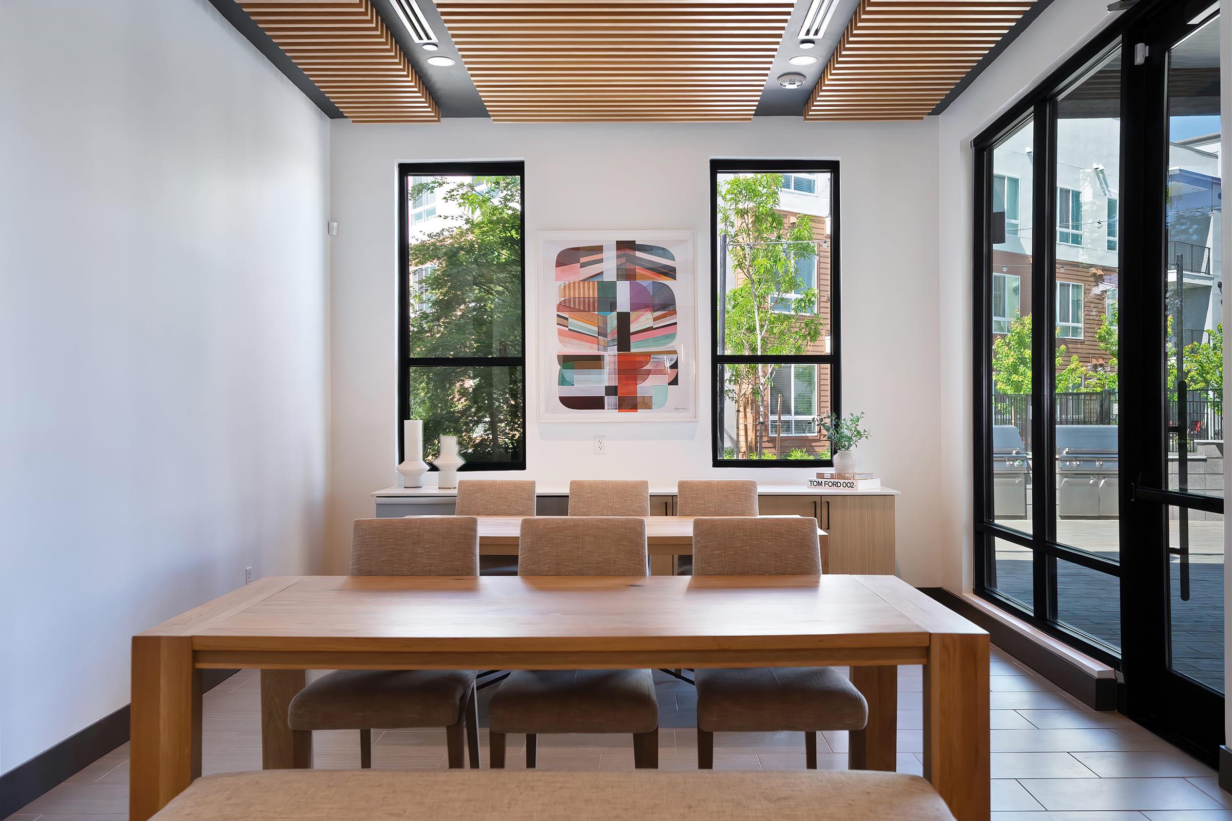 A modern dining area featuring a long wooden table with beige upholstered chairs. Large windows allow natural light, revealing greenery outside. A piece of abstract art hangs on the wall, and there are minimalist decor elements on the sideboard, contributing to the contemporary aesthetic of the space.