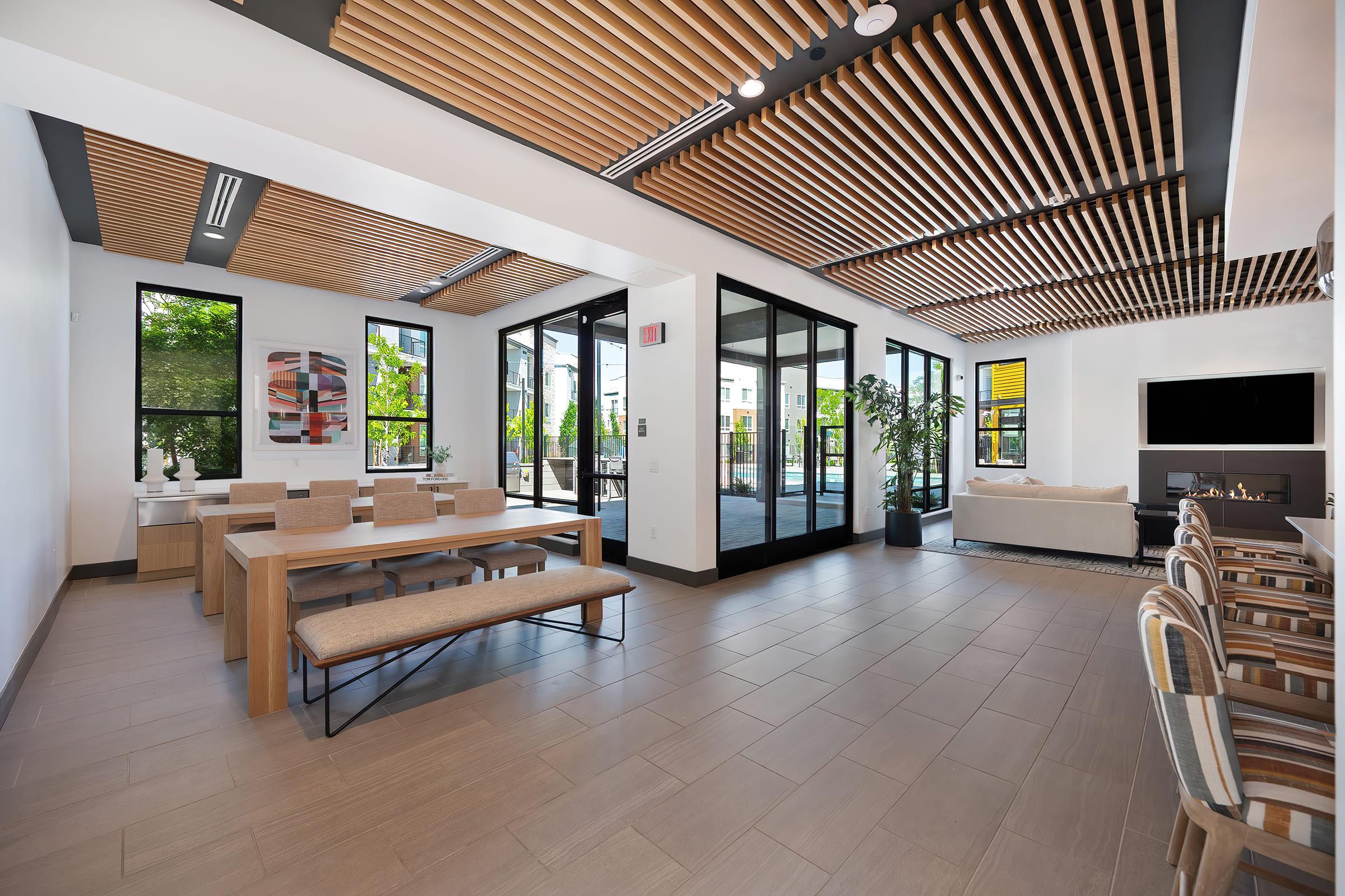 A modern interior space featuring a dining area with wooden tables and benches, large windows allowing natural light, and a cozy living area with a sofa. The ceiling is adorned with wooden beams, and there are decorative plants and artwork enhancing the aesthetic. A sliding glass door leads to an outdoor area.