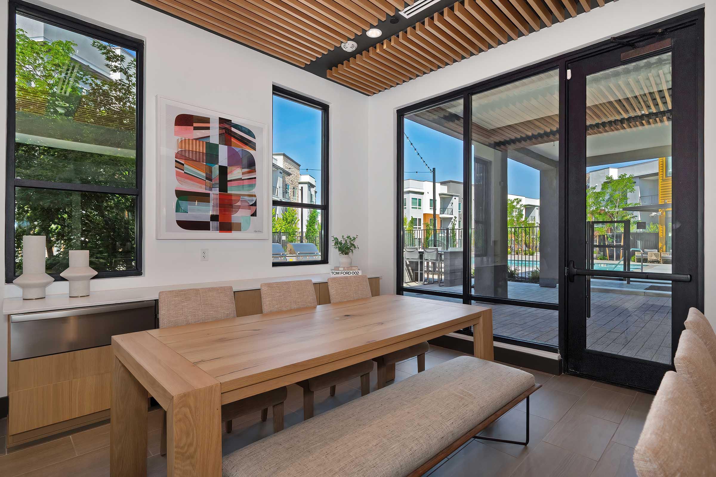 A modern dining area featuring a wooden table with beige seating, large windows providing natural light, and minimalist decor including a colorful abstract artwork on the wall. The space opens to an outdoor view with greenery and a pool area beyond the glass doors.