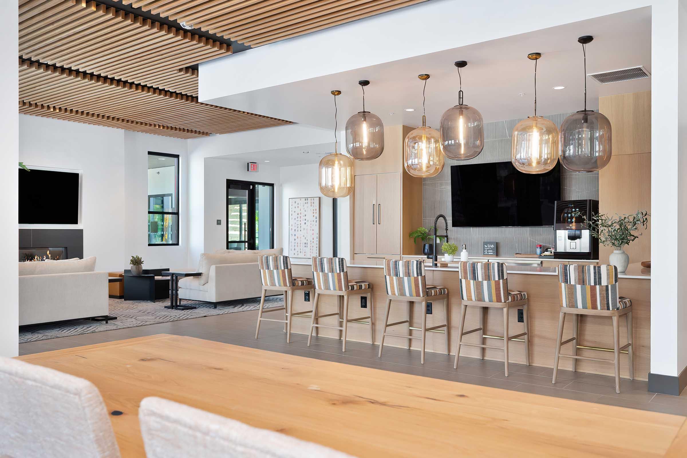 Modern kitchen and dining area featuring a large wooden table, stylish bar stools, and pendant lighting. The kitchen has a sleek design with wooden cabinetry and a black countertop. A comfortable seating area with sofas is visible in the background, along with large windows allowing natural light.