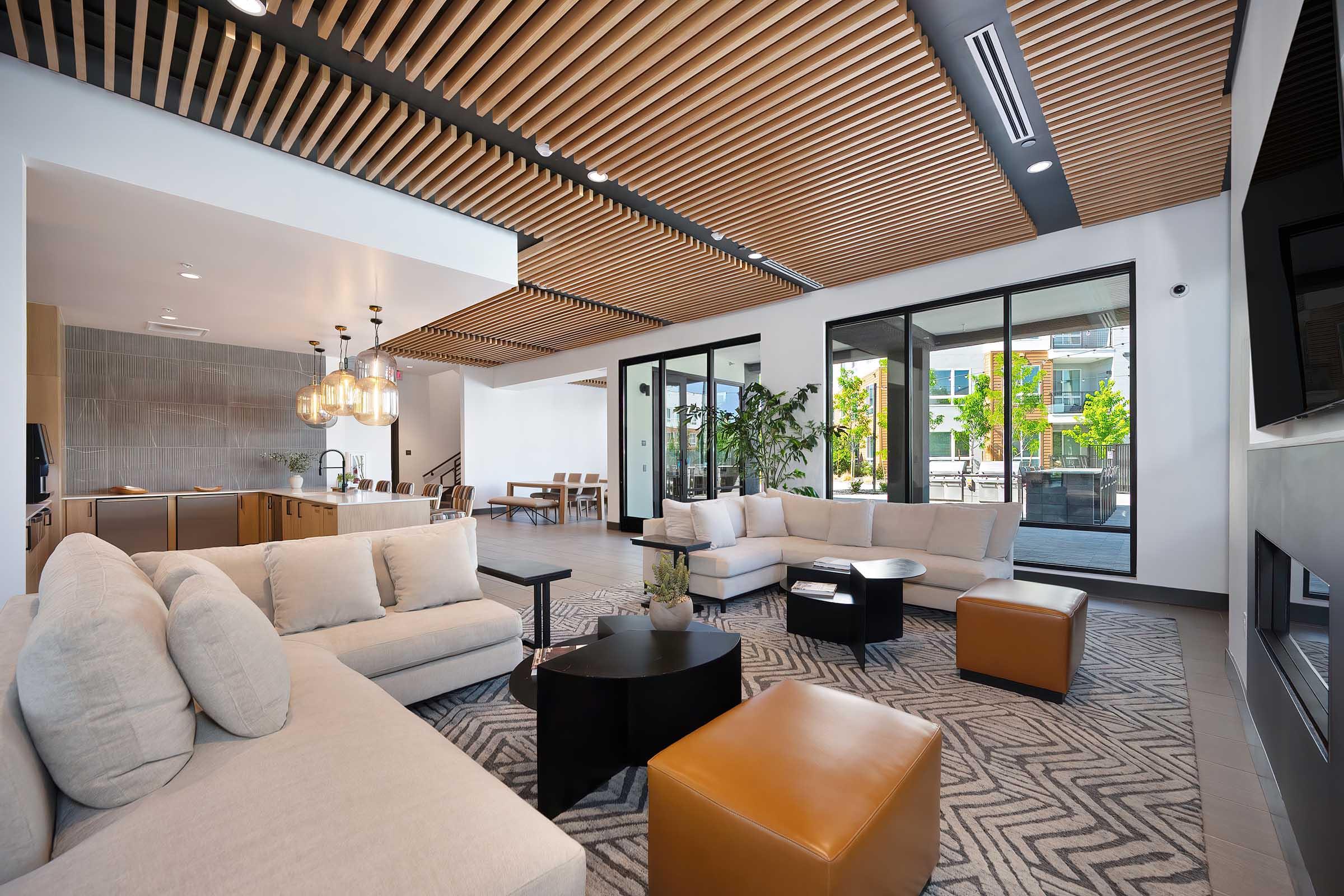 A modern, spacious living area featuring a stylish arrangement of beige sofas, a dark wood coffee table, and an ottoman. Bright natural light pours in through large glass doors, revealing greenery outside. The ceiling showcases a unique wooden slat design, adding warmth to the contemporary decor.