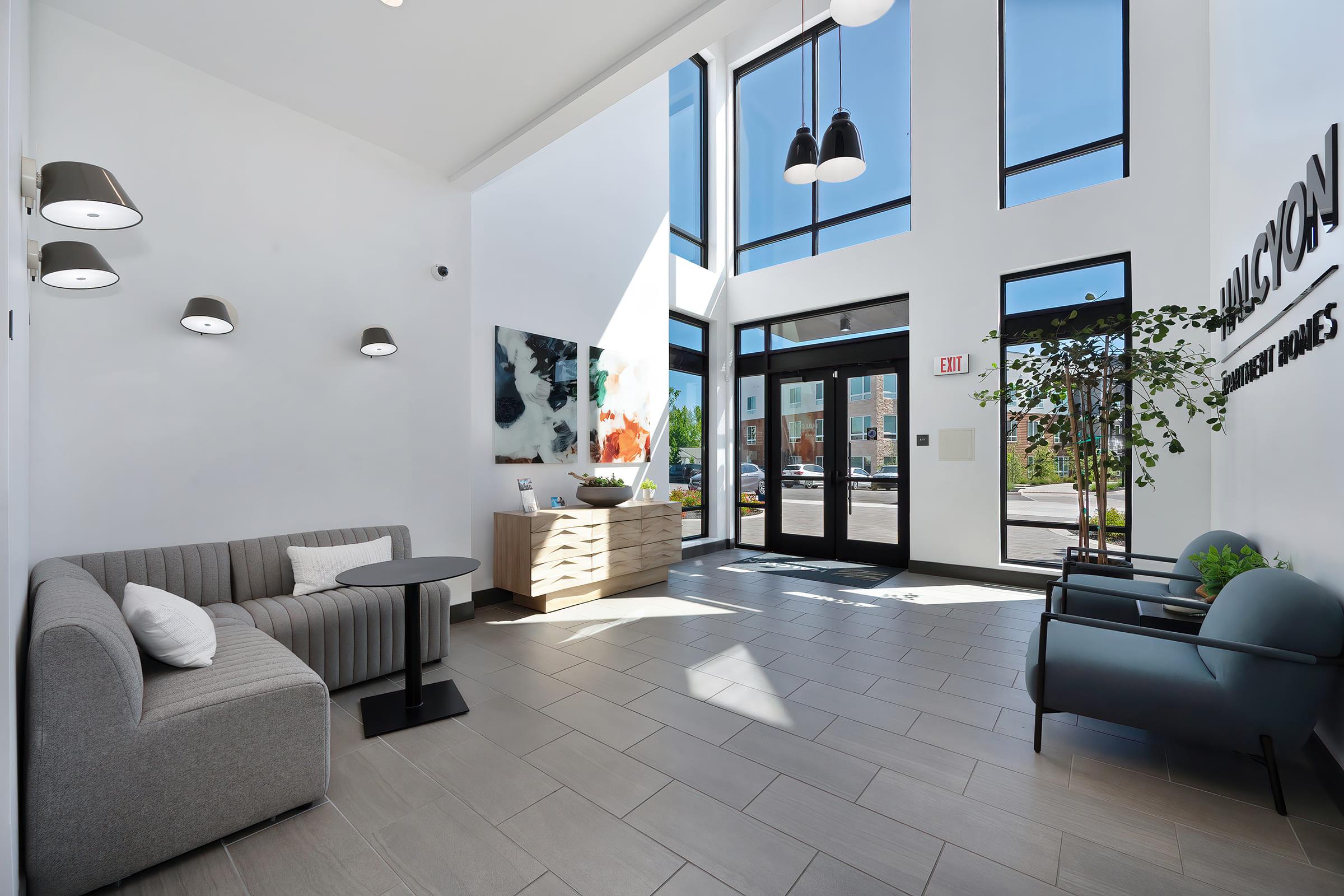 A modern lobby featuring large windows that let in natural light. There is a gray sofa and a small round table on one side, while on the opposite side, there are two black chairs. A wooden cabinet holds decorative items, and a colorful piece of art is displayed on the wall. Bright and inviting atmosphere.