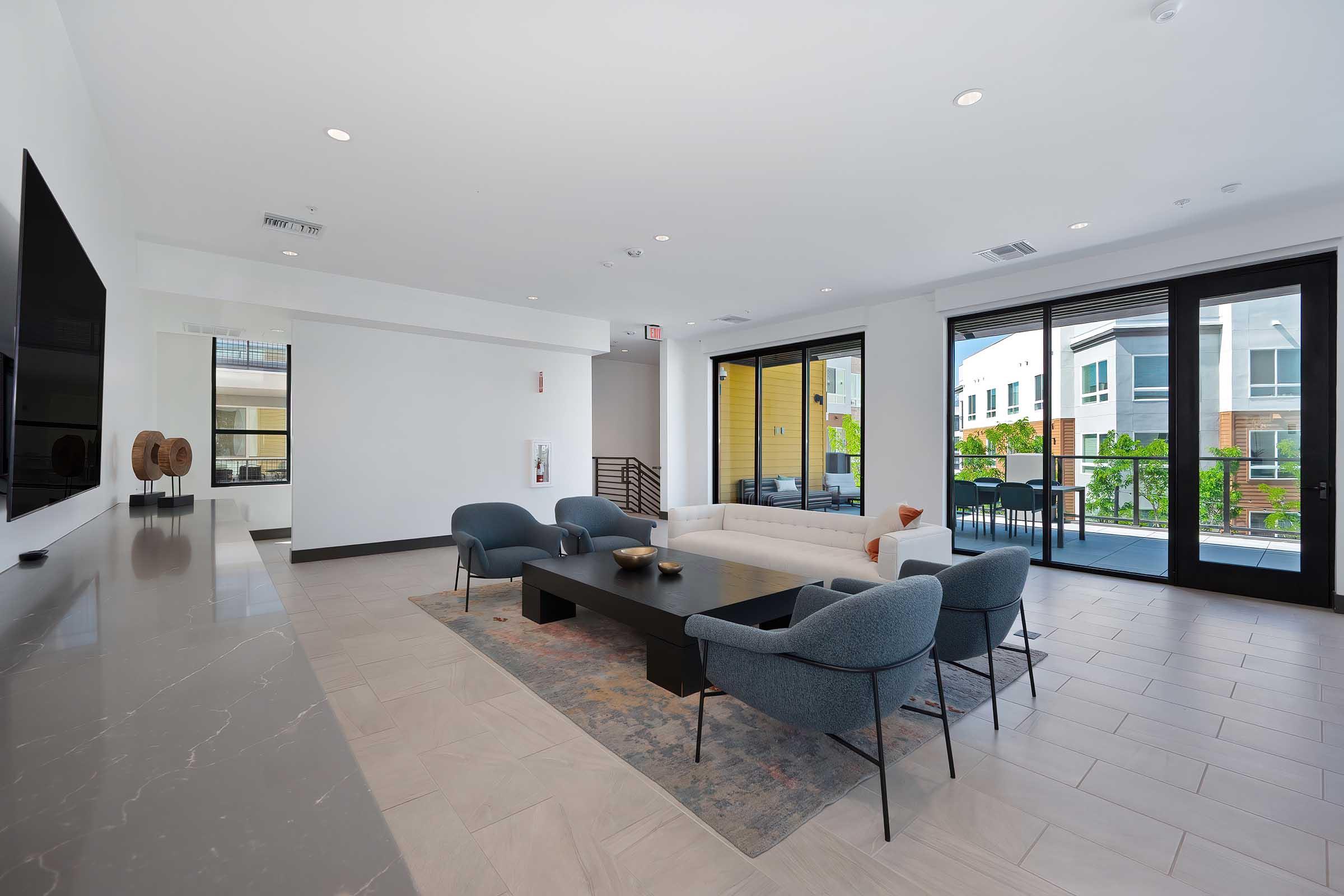 A modern living room with a light color scheme, featuring a white sofa, dark coffee table, and four blue armchairs. Large windows allow natural light, revealing a view of an outdoor area. The floor is tiled, and there is a flat-screen TV mounted on the wall, complemented by decorative elements.