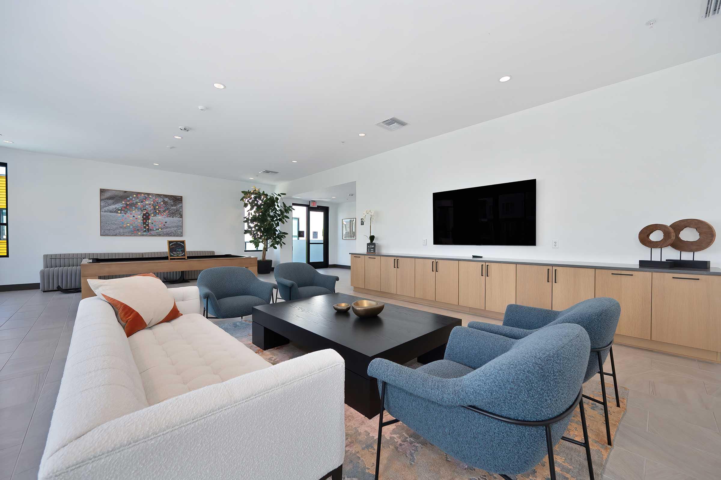 A modern living room featuring a light-colored sofa, blue accent chairs, and a dark coffee table. The walls are painted white, and large windows allow natural light. A flat-screen TV is mounted on the wall, and a piece of abstract art is displayed. Decorative items are arranged on the furniture.