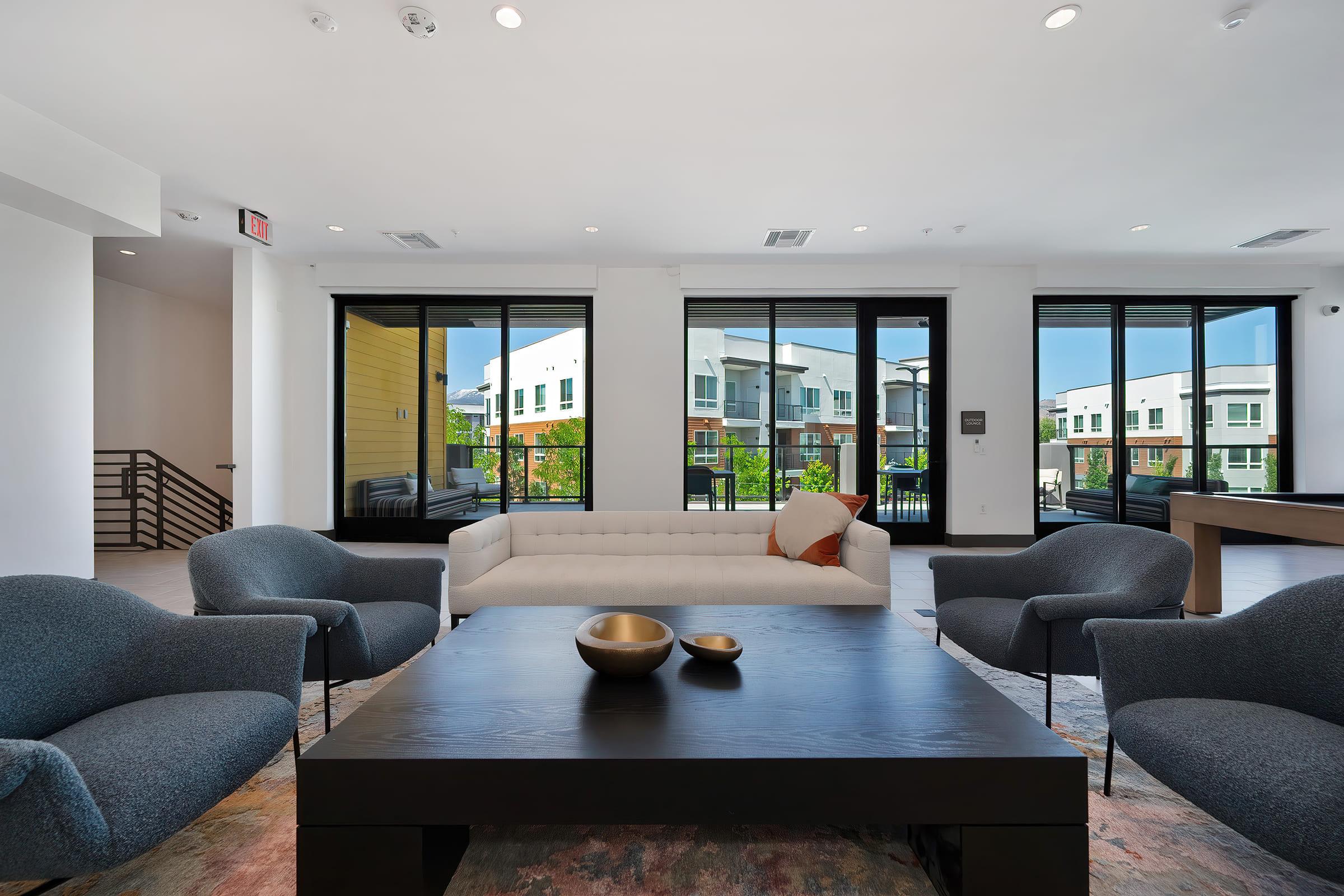 A modern living room featuring a sleek coffee table with a bowl, a light-colored sofa, and two gray armchairs. Large windows provide a view of an outdoor area with buildings and greenery. The decor is minimalist and contemporary, creating a bright and inviting atmosphere.
