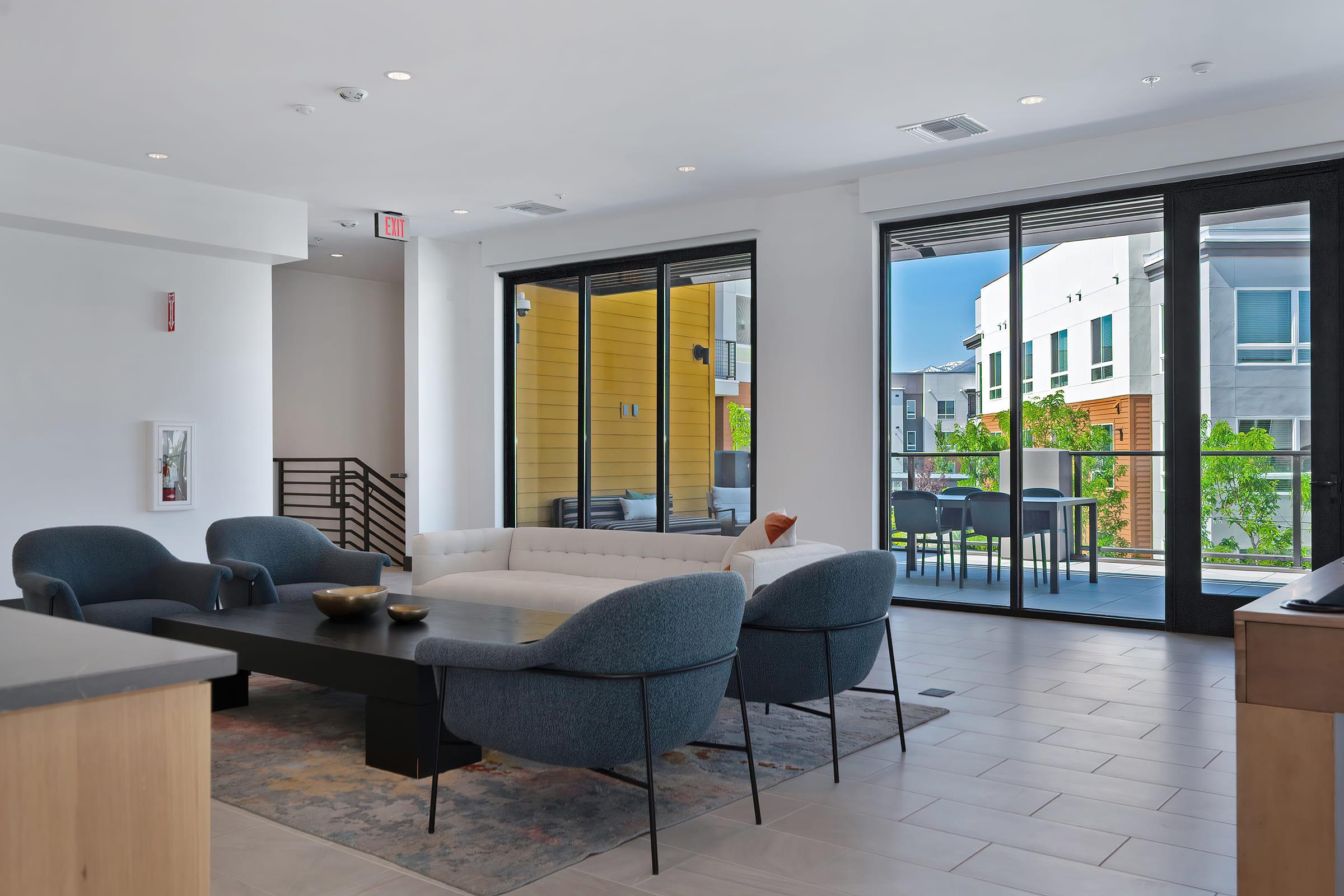 Modern living room with sleek furniture, including a light-colored sofa, blue chairs, and a dark coffee table. Large windows allow natural light to enter, showcasing an outdoor balcony with greenery. The space features a minimalist design with neutral colors and contemporary decor.