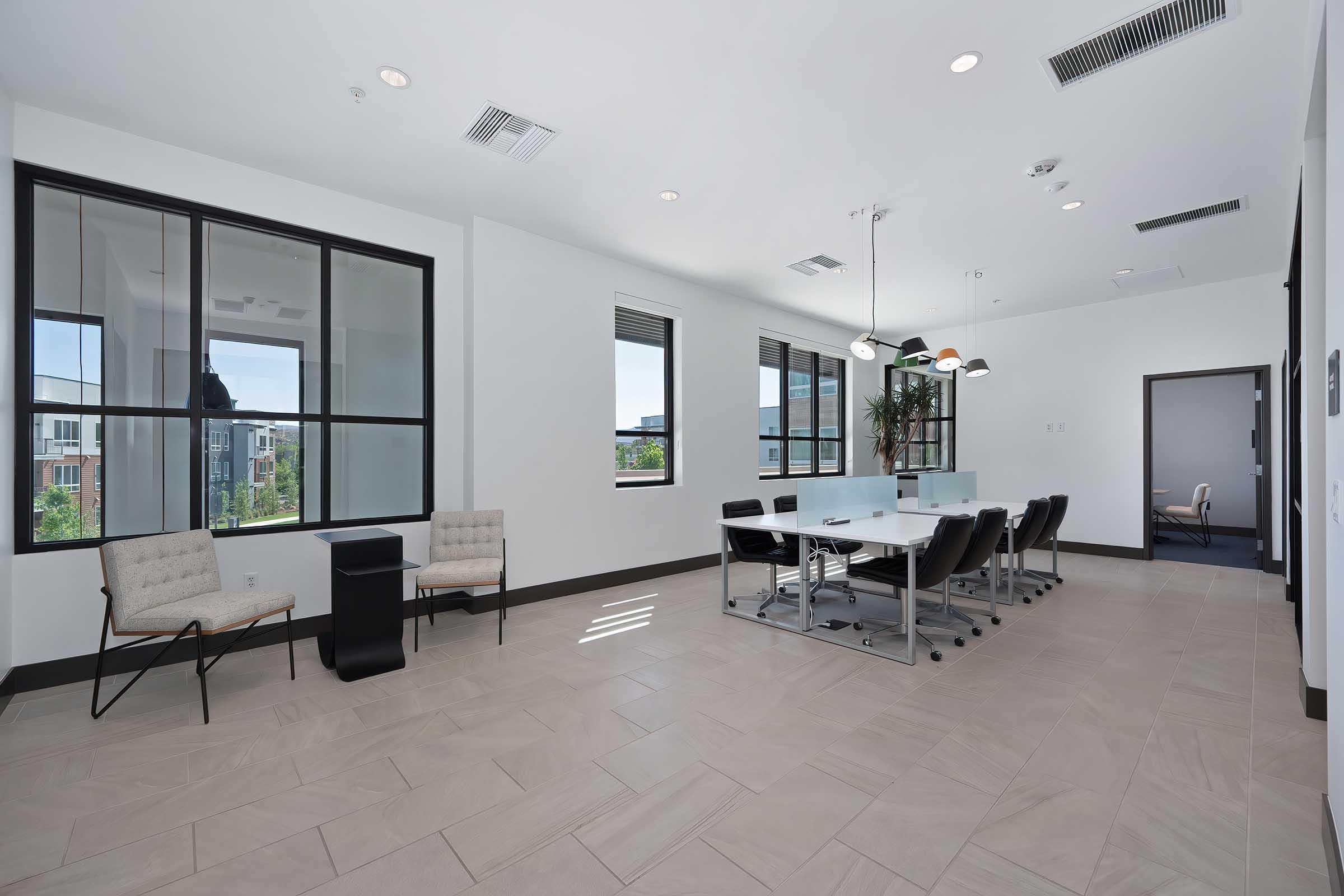 A modern conference room featuring a long table with chairs, large windows allowing natural light, and minimalistic decor. Two beige chairs are positioned near a small black side table. The room has a bright and spacious feel, ideal for meetings or collaborative work.
