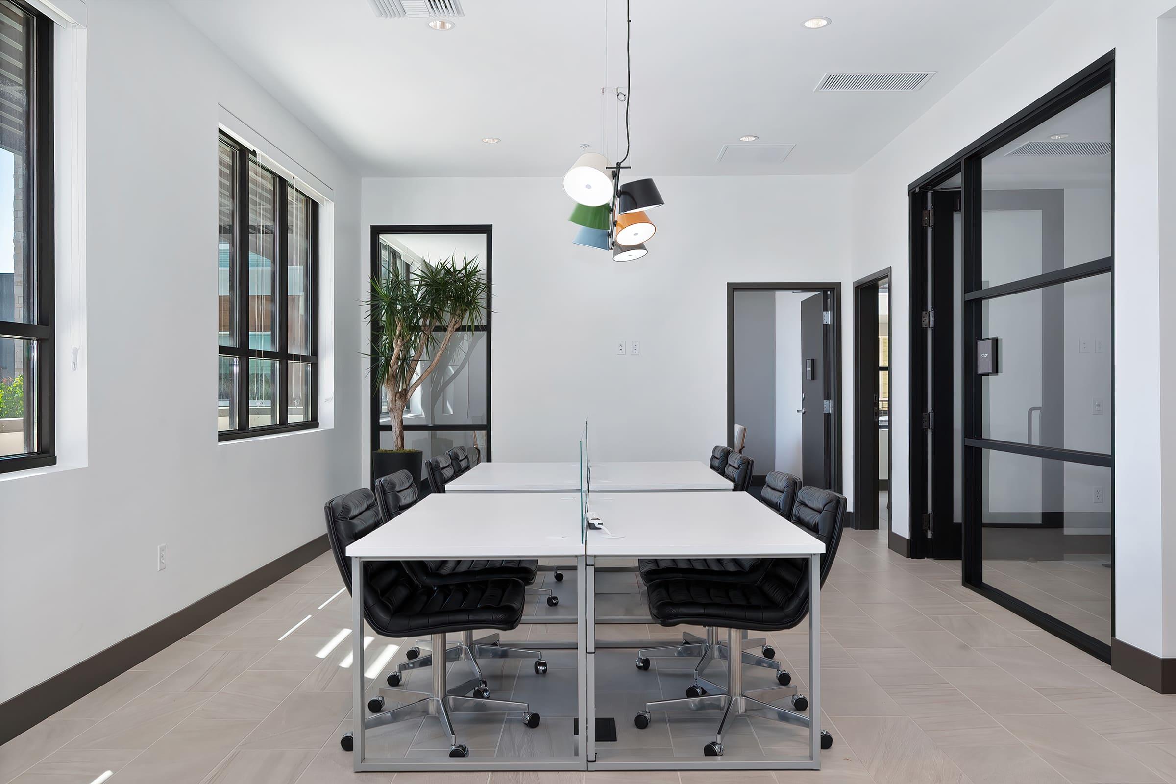 A modern conference room featuring a large white table with black ergonomic chairs, large windows letting in natural light, a stylish pendant light fixture, and a potted plant in the corner. The space is designed with a minimalist aesthetic and has glass doors leading to another area.