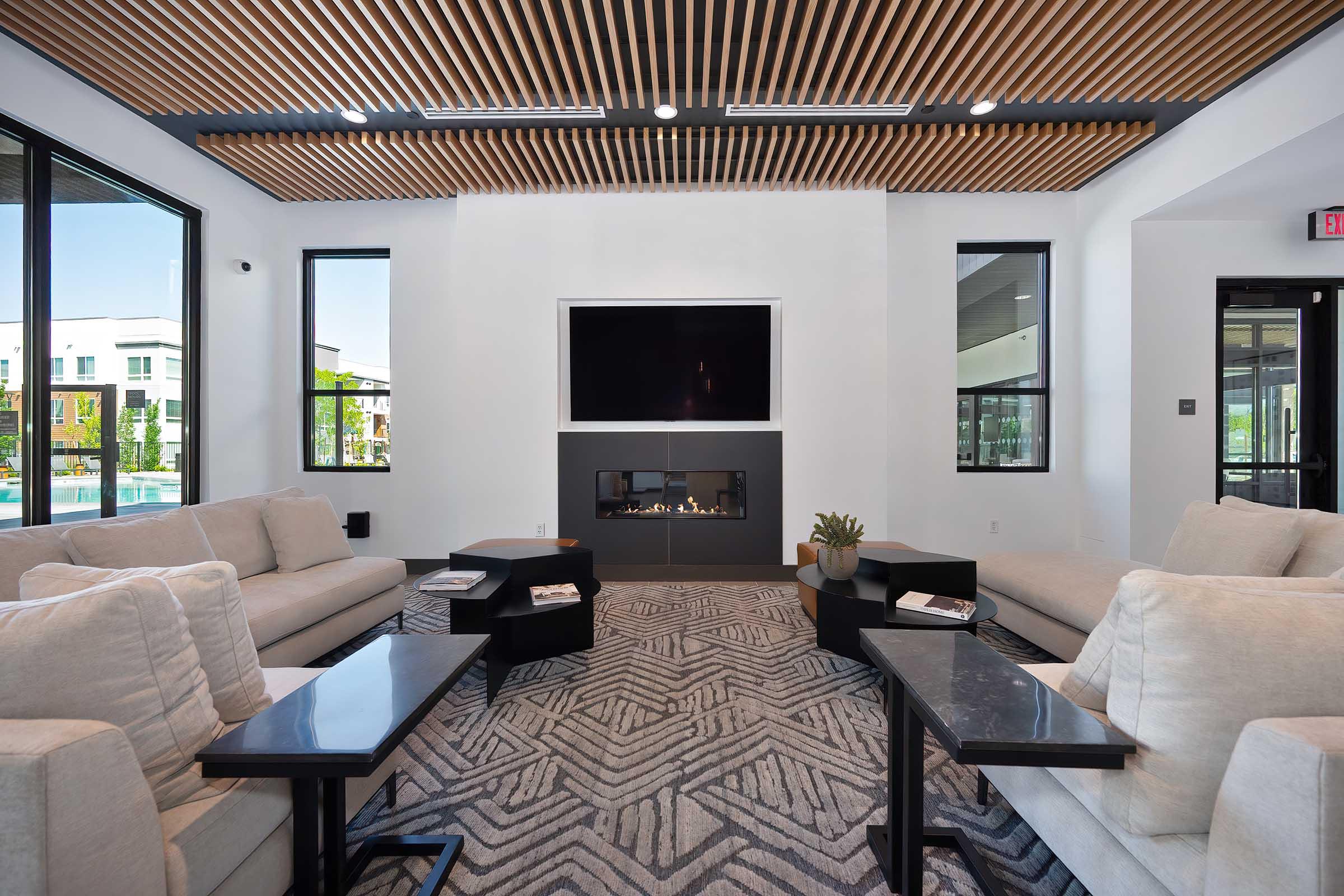 A modern lounge area featuring two light-colored sofas facing a sleek fireplace. There are small, dark side tables with magazines on them. A large flat-screen TV is mounted on the wall above the fireplace. The space is illuminated by natural light coming through large windows, with a patterned rug on the floor.