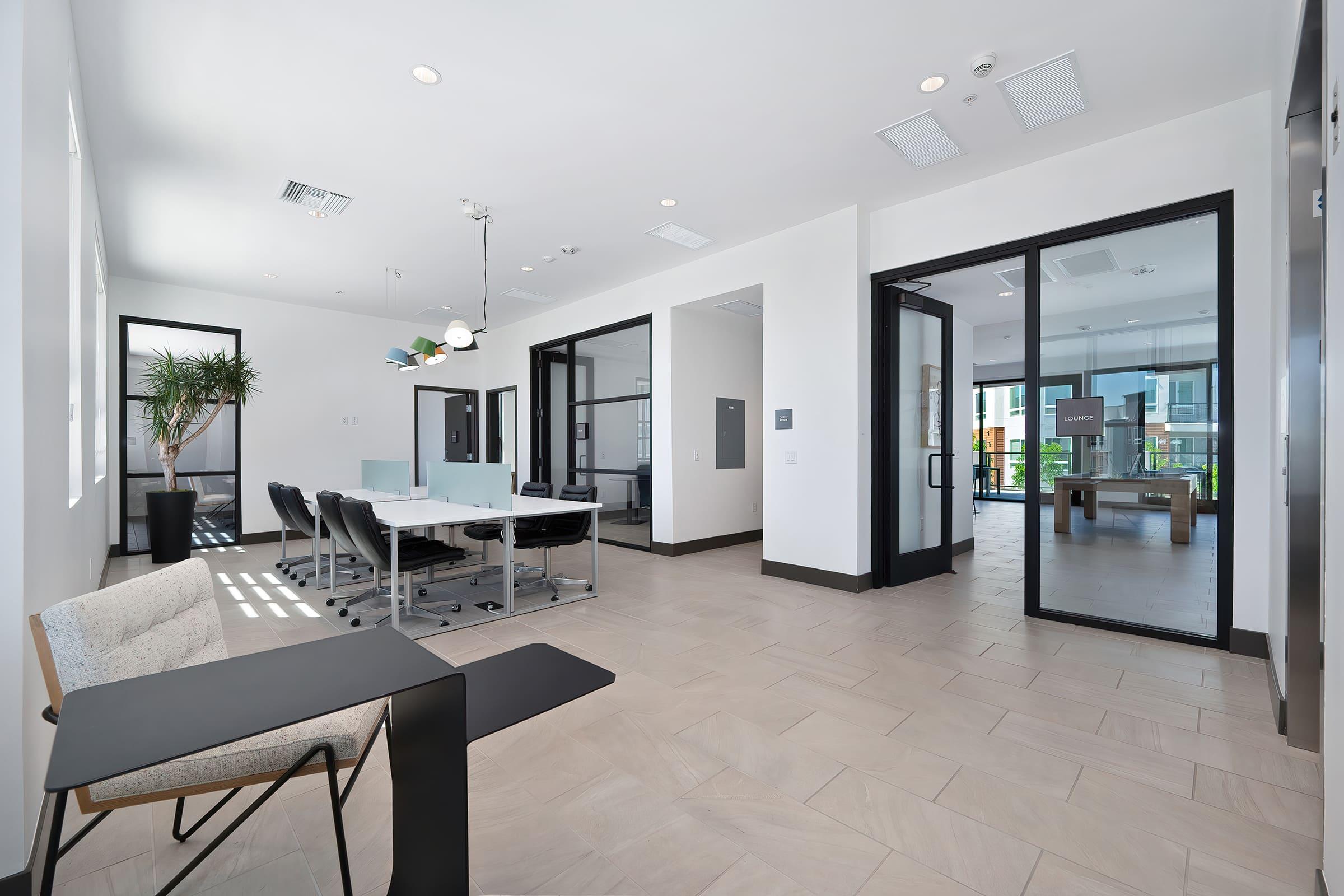 Modern office space featuring a conference room with a glass table and black chairs, natural light streaming through large windows. A minimalist lounge area is visible with a small table and chair, along with indoor plants adding a touch of greenery. Sleek, contemporary decor throughout the space.