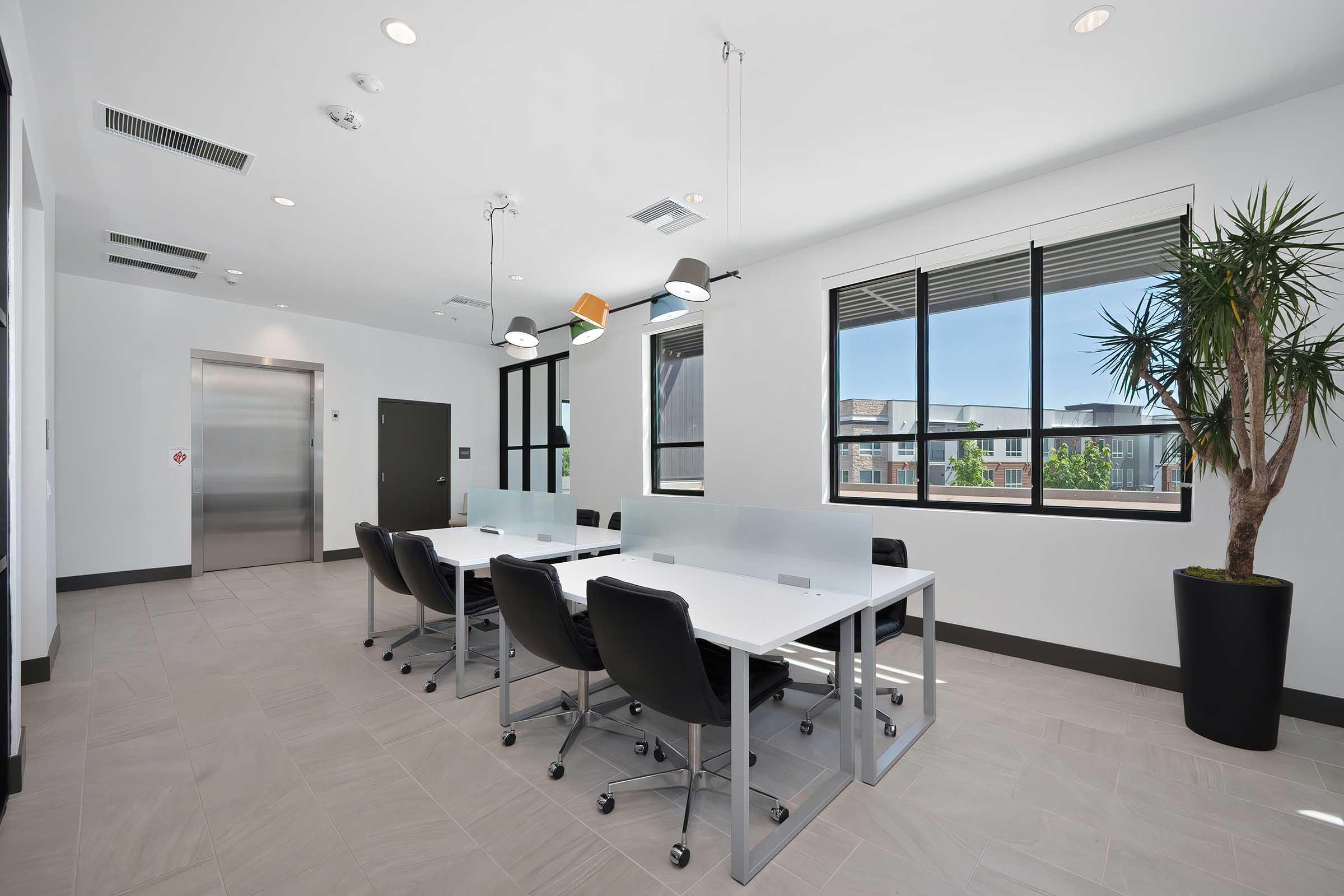 Modern conference room featuring a large white table surrounded by black office chairs, large windows allowing natural light, a vertical indoor plant in the corner, and an elevator door visible. Bright lighting fixtures hang from the ceiling, creating a professional and airy atmosphere.