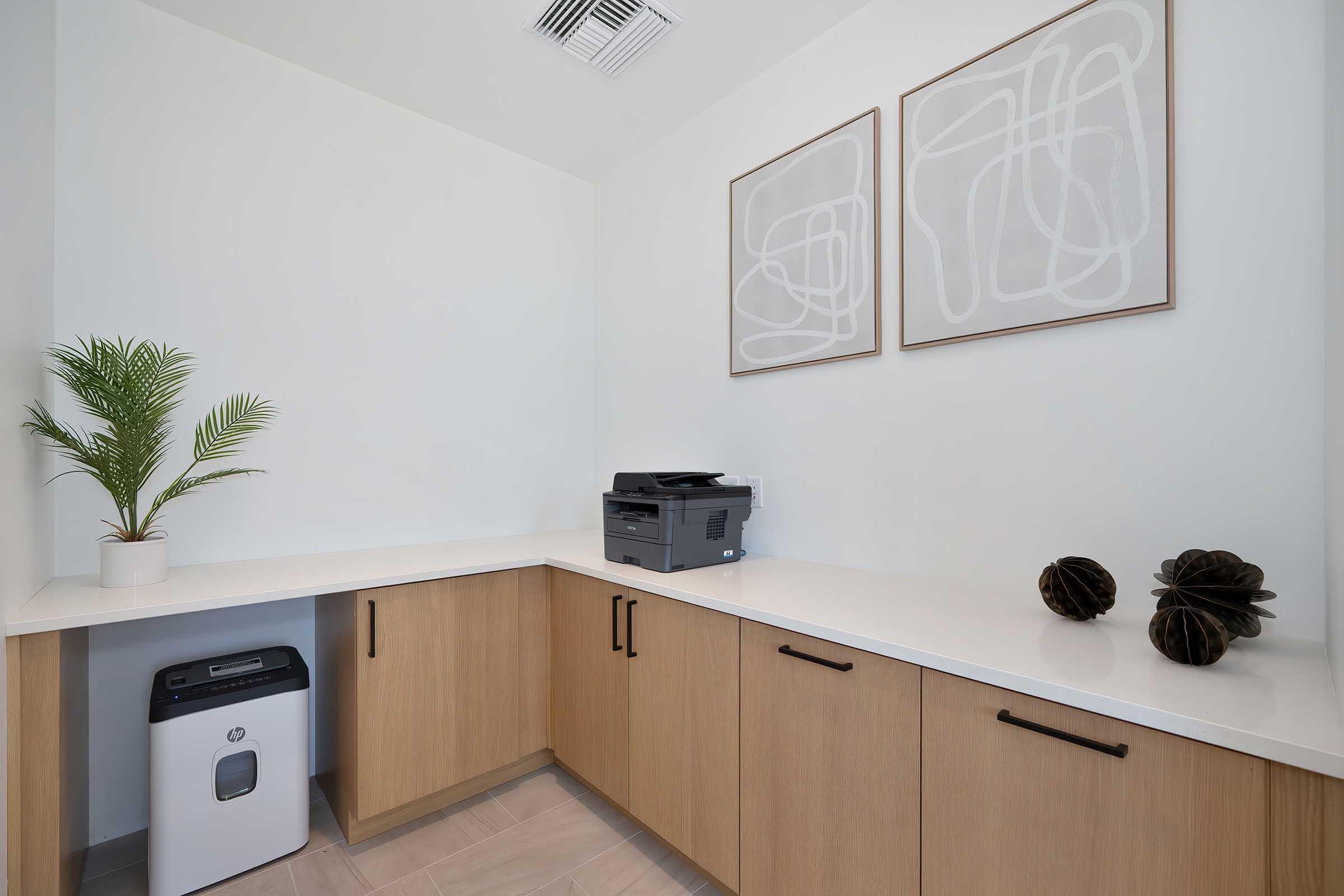 A modern office space featuring a light wooden countertop with a printer, a small trash bin, and decorative elements like a potted plant and two abstract framed artworks on the wall. The design is minimalist and clean, with neutral tones dominating the color palette.