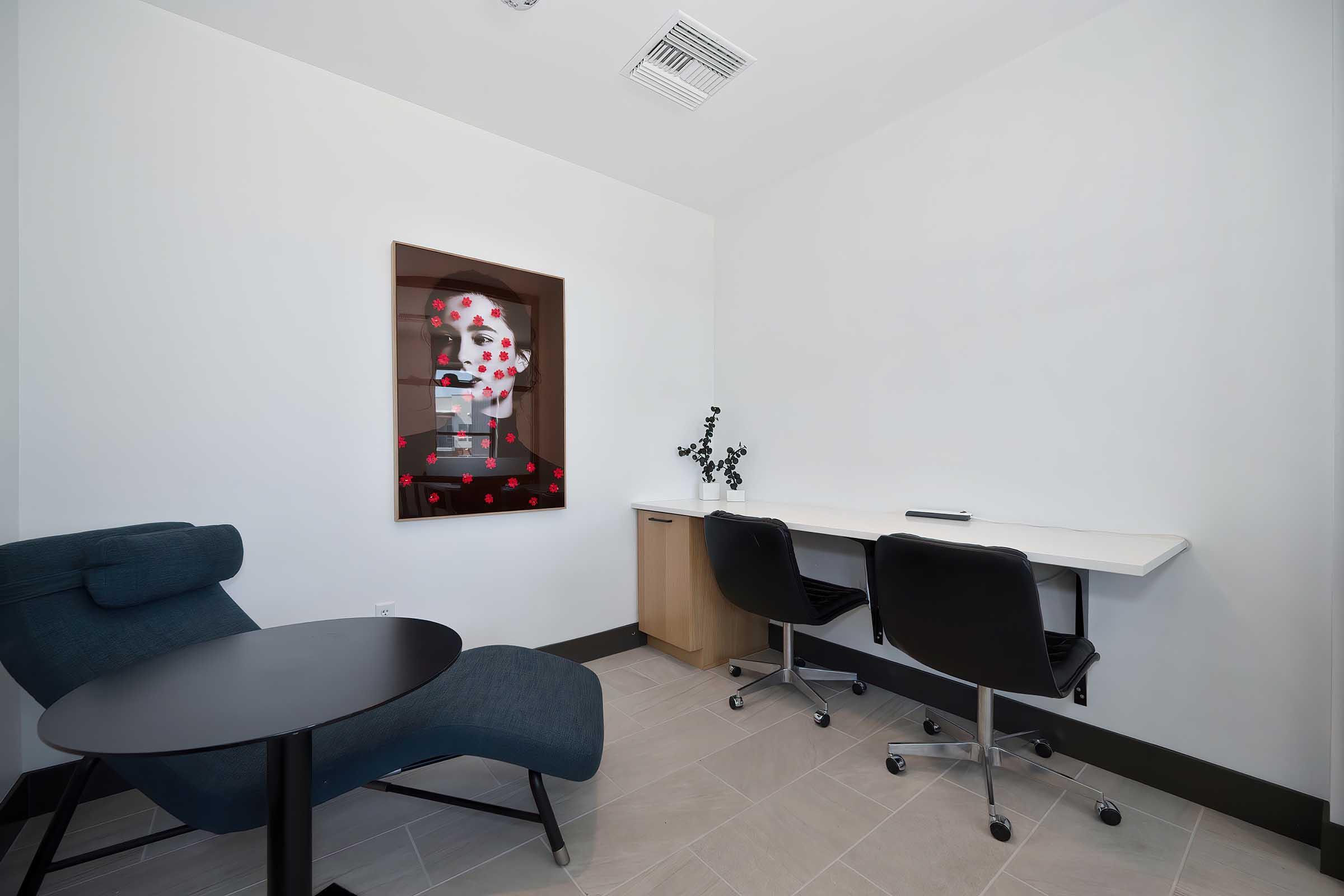 A modern interior of a small room featuring a blue lounge chair, a black round table, and two black office chairs next to a white desk. On the wall, there is a framed artwork with a face surrounded by red dots. The walls are painted white, and there is a small plant on the desk.