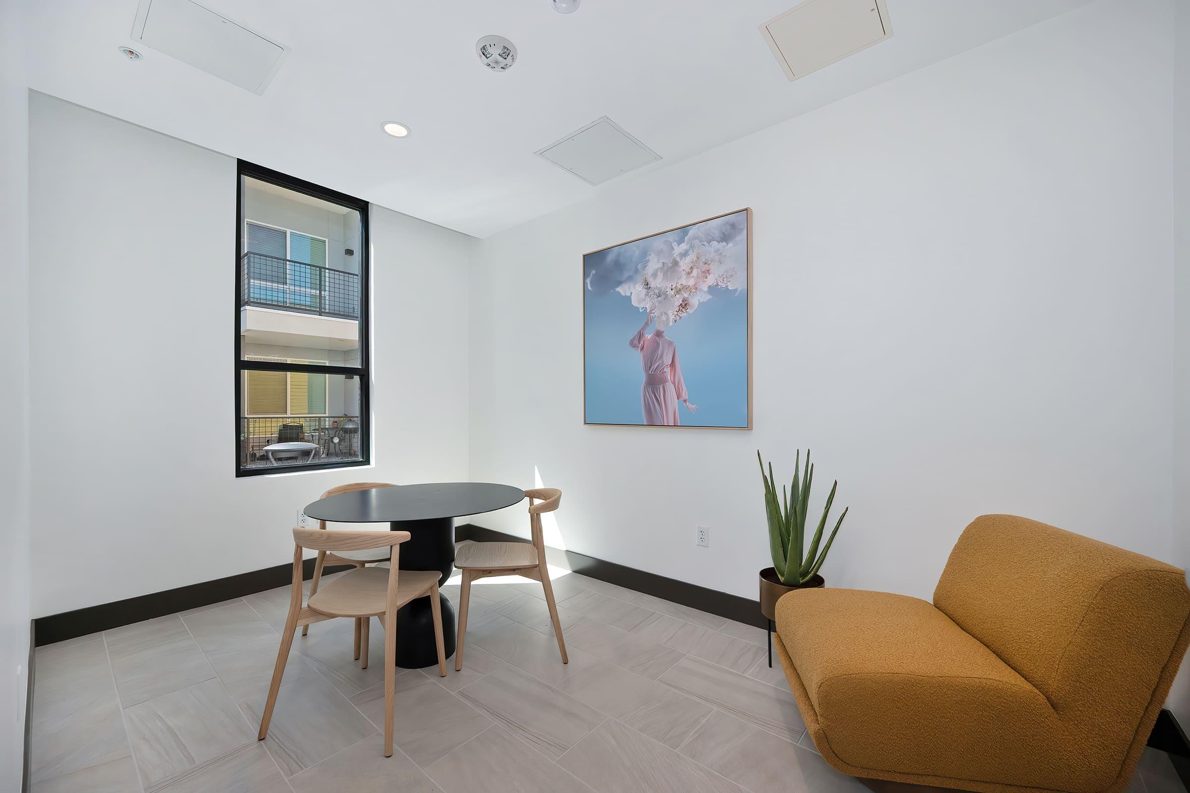 A modern interior room featuring a round black table with two wooden chairs, a cozy mustard-colored armchair, and a large artwork on the wall depicting a figure with a cloud above their head. A green plant is in one corner, and a window allows natural light into the space.