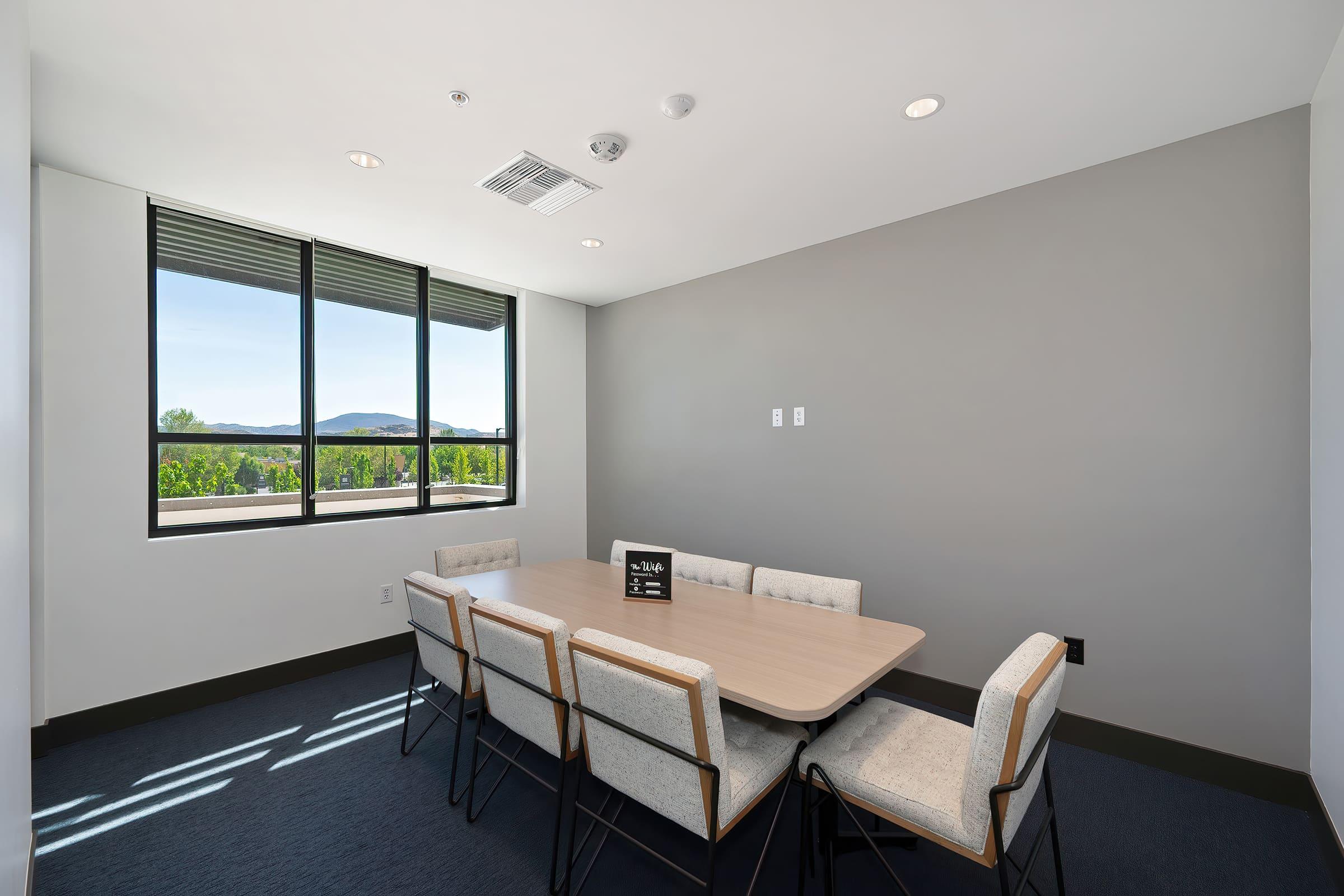 A modern meeting room featuring a rectangular wooden table surrounded by six light-colored chairs. Large windows provide natural light and a view of greenery outside. The walls are painted in neutral tones, and there is a ceiling with recessed lighting.