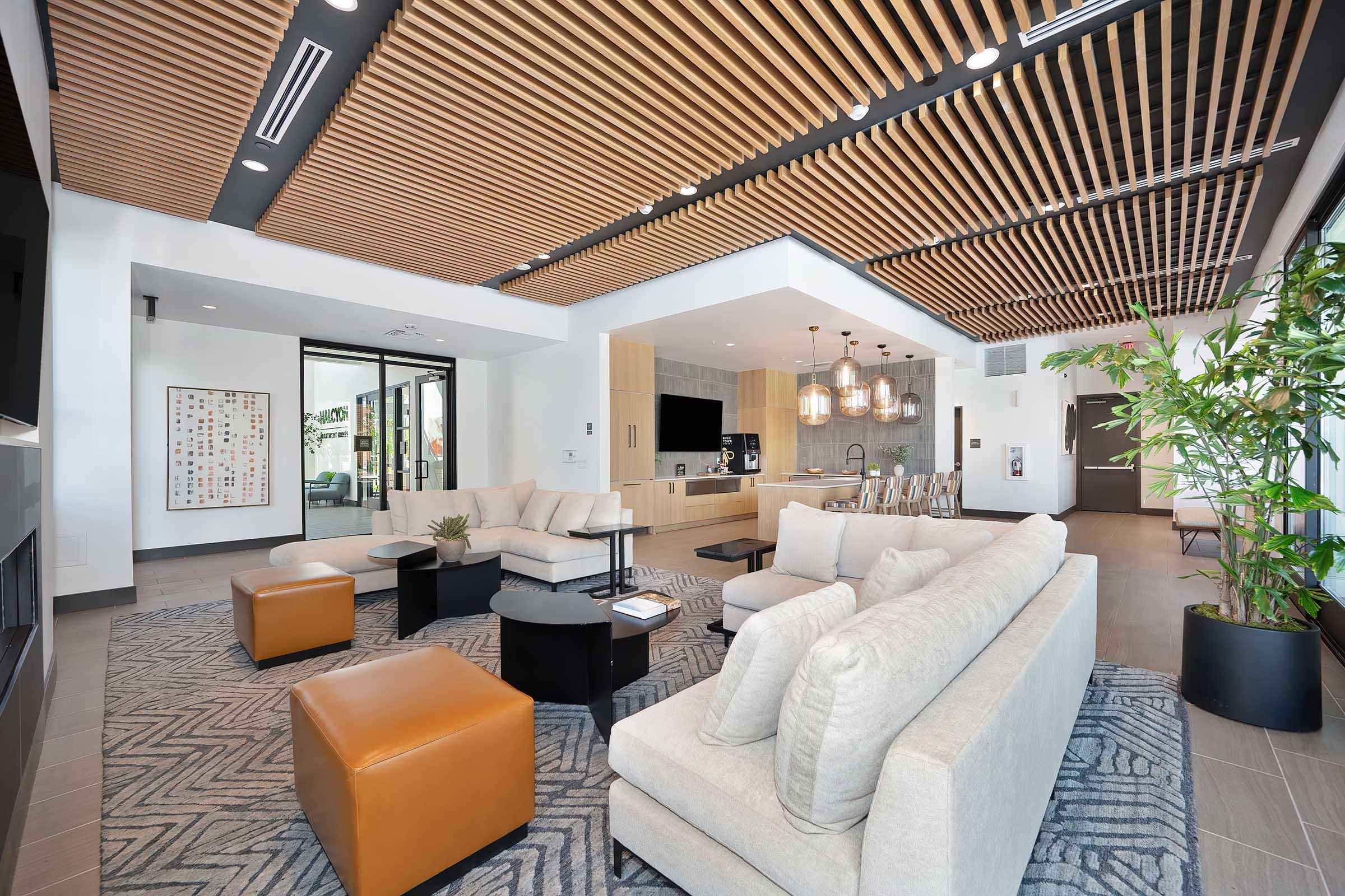 Modern living room and kitchen area in an upscale apartment. Features comfortable beige sofas, stylish armchairs, and a sleek coffee table. The ceiling is adorned with wooden slats, and large windows allow natural light. Decor includes a plant and a contemporary artwork piece on the wall.