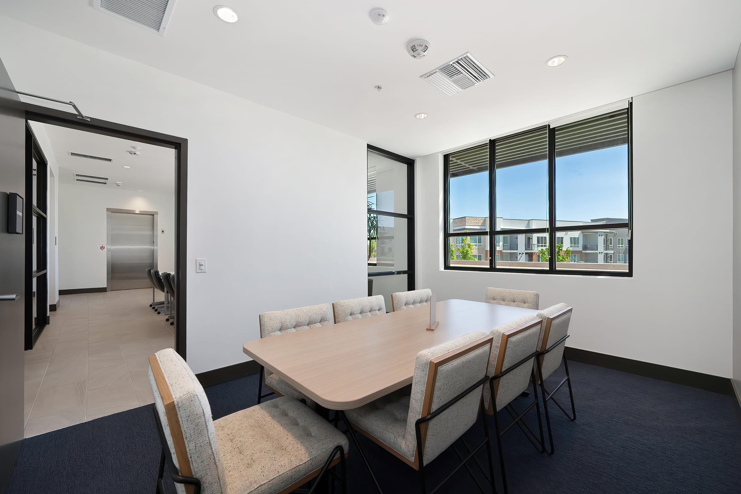 A modern conference room featuring a rectangular wooden table surrounded by upholstered chairs. Large windows allow natural light to flood the space, with views of greenery outside. The walls are painted white, and the flooring is dark, creating a contemporary and professional atmosphere.