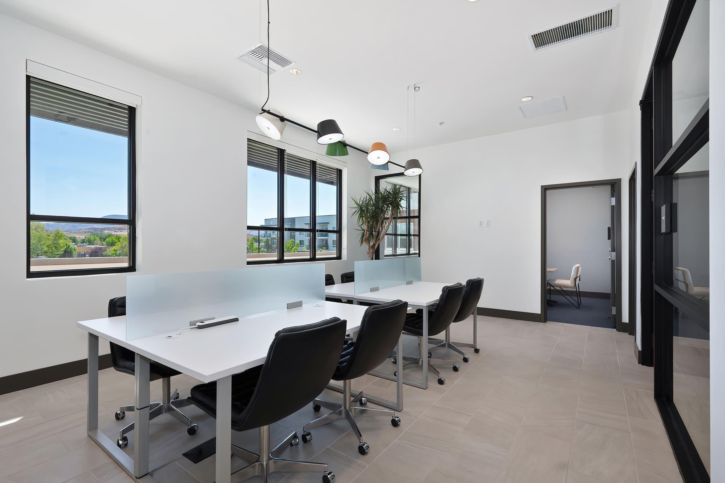 Modern conference room featuring large windows with natural light, sleek white tables, and black rolling chairs. There are contemporary pendant lights hanging from the ceiling and a potted plant near the window. An adjoining door and a light-colored wall complete the minimalist design.