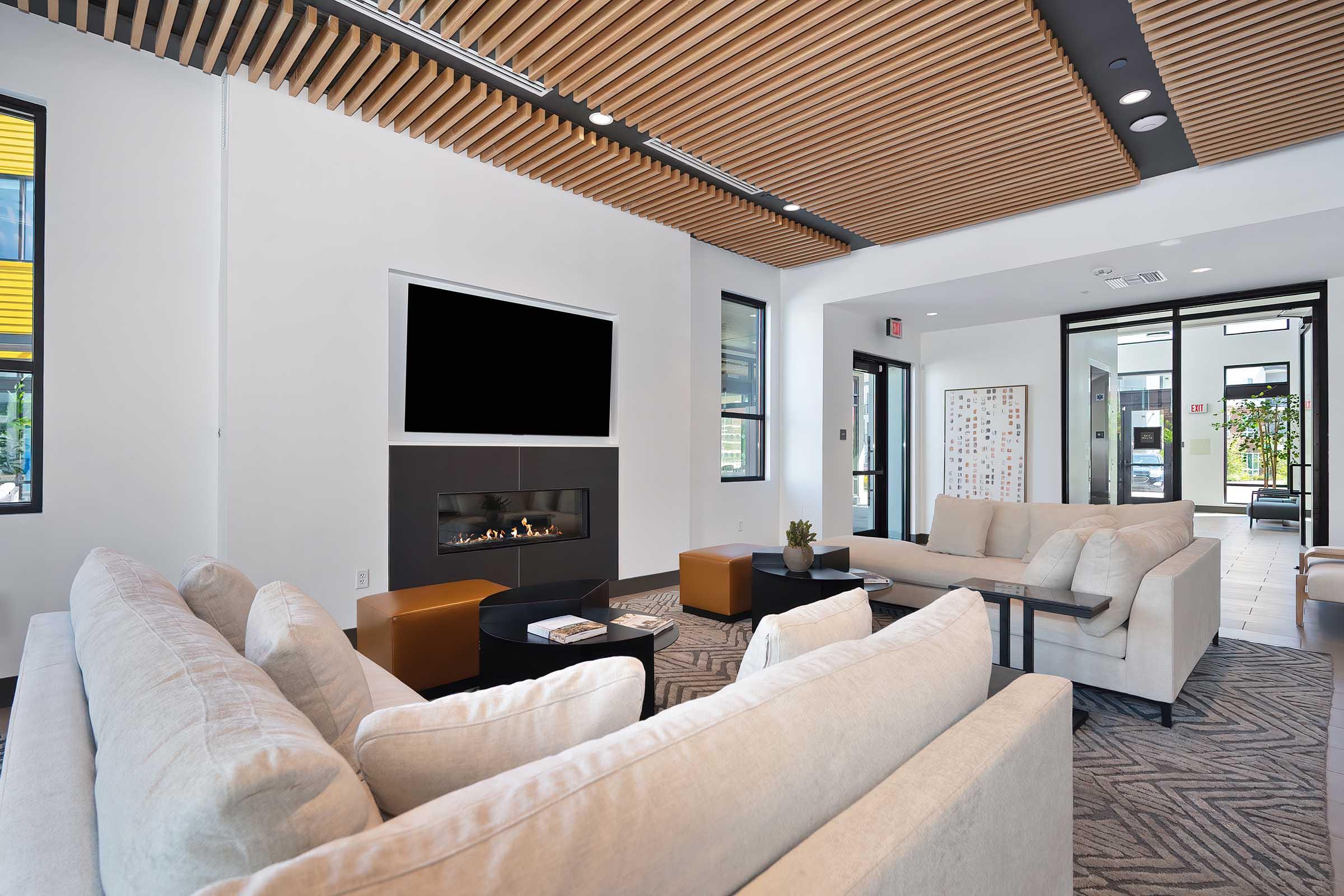 A modern lounge area featuring two plush beige sofas, a coffee table, and an elegant fireplace. The space has large windows allowing natural light, a stylish wooden ceiling, and comfortable seating. There is a small plant on the table and a welcoming atmosphere with contemporary decor.