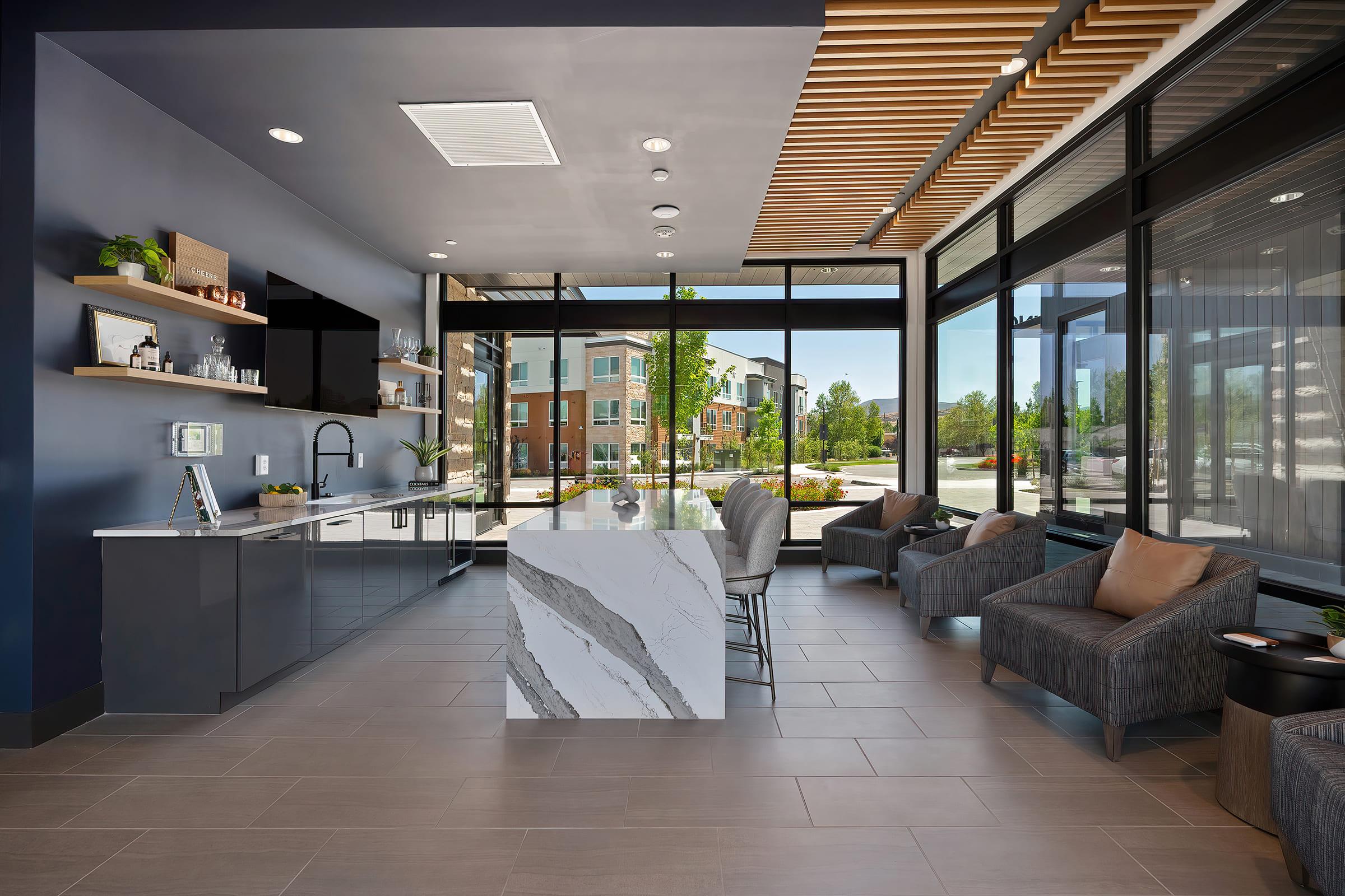 Modern lobby area featuring a sleek kitchen island with a marble top, high-backed chairs, and large windows offering a view of the outdoors. Shelves with plants and decor enhance the contemporary design, while neutral-colored walls and flooring create a welcoming atmosphere.