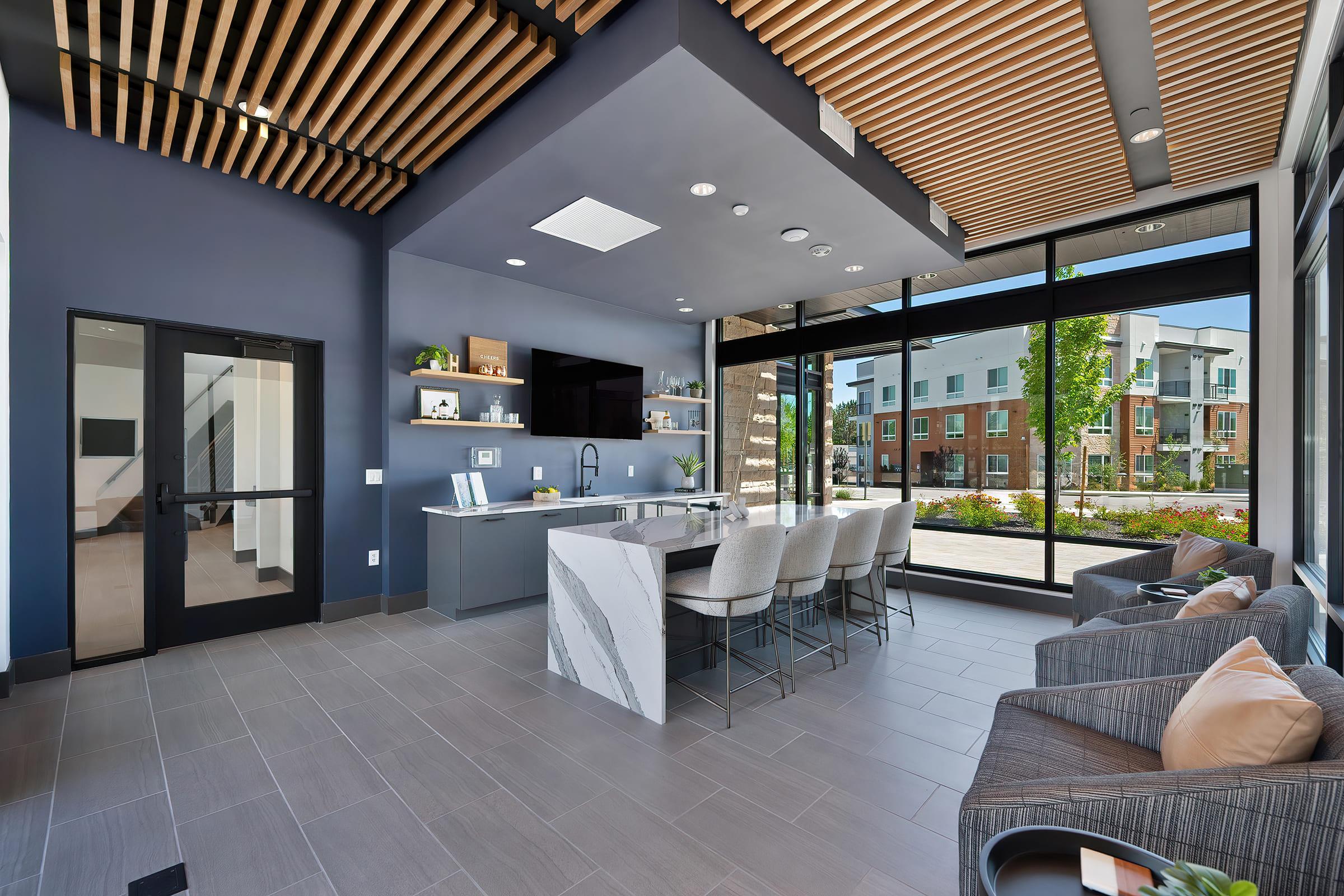 Modern lounge area featuring sleek furniture, large windows with outdoor views, a stylish kitchen island, and a cozy seating arrangement. The room has a contemporary design with wooden ceiling elements and neutral tones, creating an inviting atmosphere.