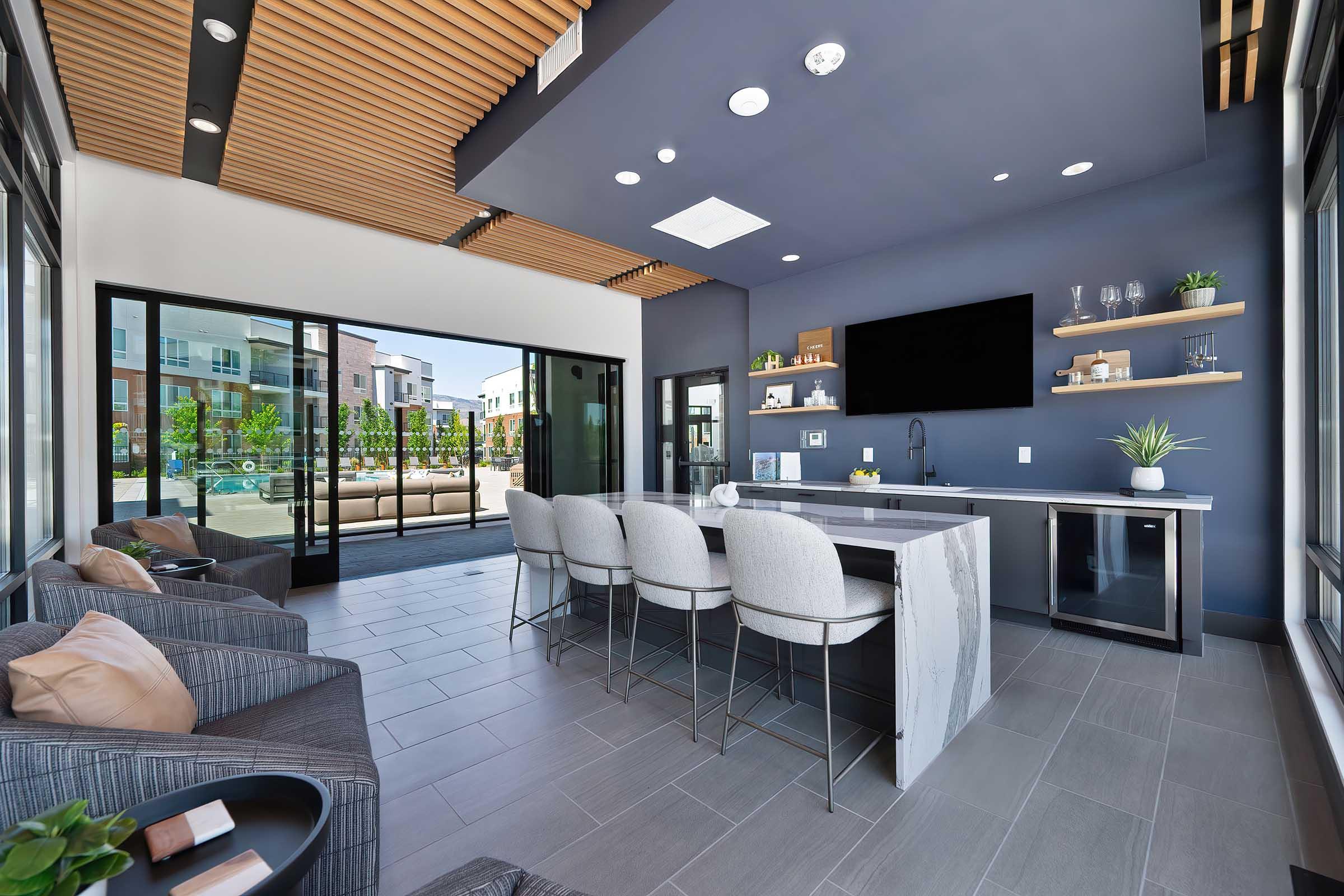 Modern lounge area featuring a stylish kitchen island with bar seating and a flat-screen TV. Large windows provide natural light and a view of an outdoor patio space. The interior design includes a mix of gray tones, wooden accents, and decorative plants for a contemporary feel.