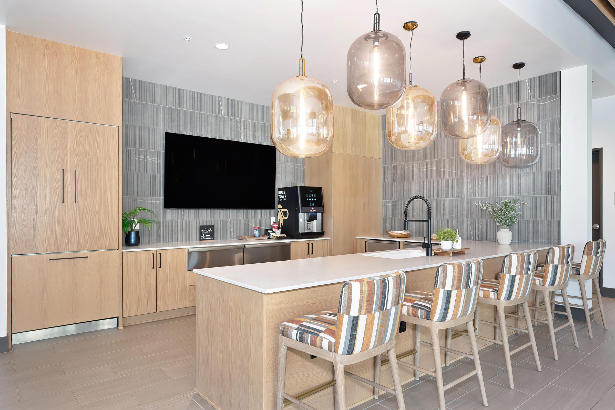 Modern kitchen interior featuring light wood cabinetry, a spacious island with bar seating, and stylish pendant lighting. A large flat-screen TV is mounted on the wall, and there is a coffee station visible. The decor is minimalistic with neutral tones and subtle greenery.