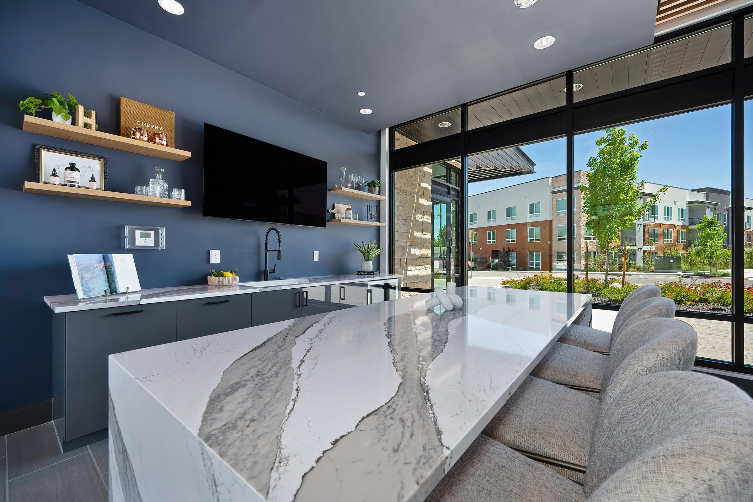 A modern kitchen interior featuring a large island with a gray and white speckled countertop. The space has sleek cabinets, a wall-mounted TV, and large windows providing views of an outdoor area with greenery and buildings in the background. The design is contemporary and inviting.