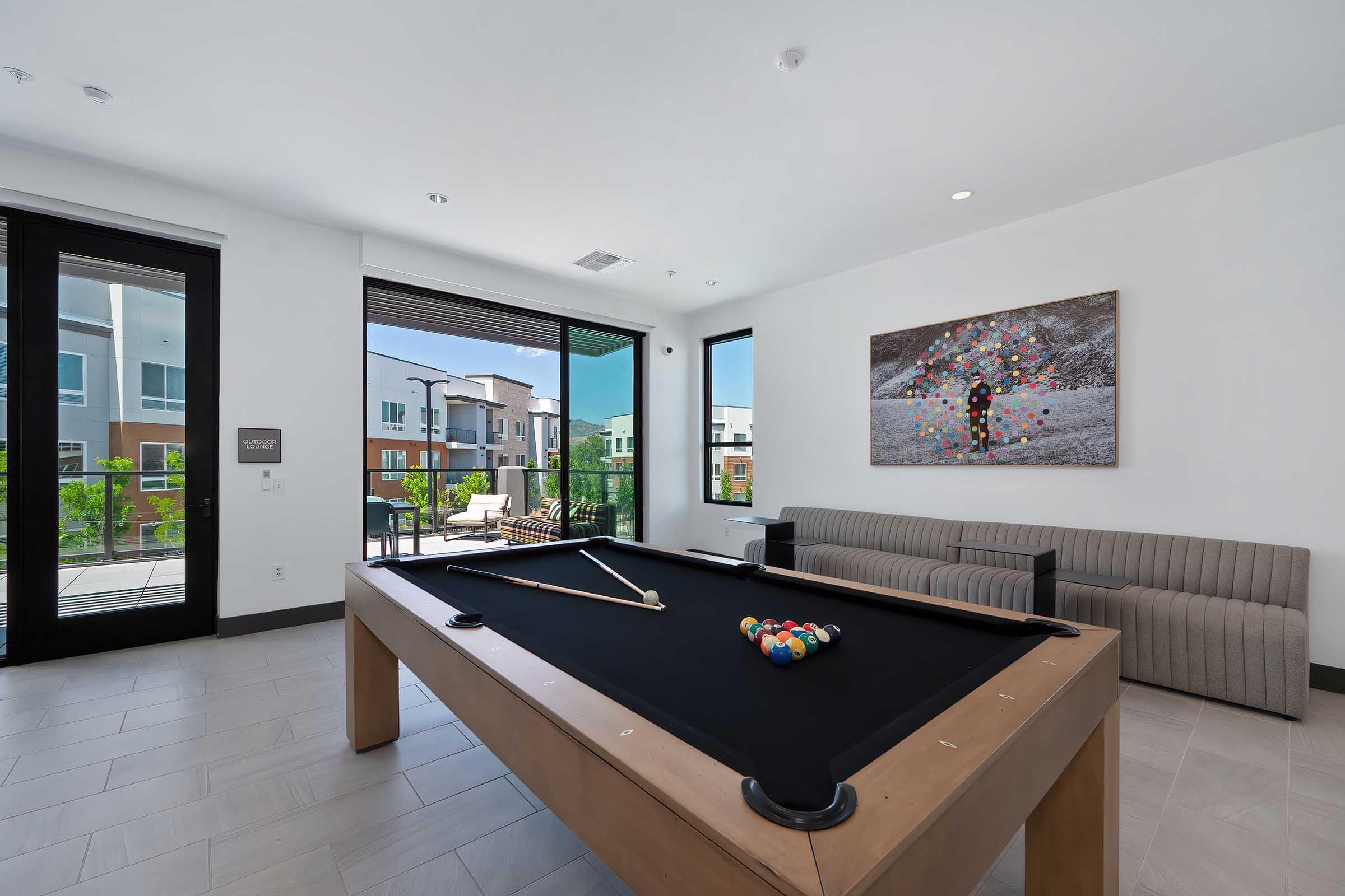 A modern indoor game room featuring a billiards table with balls arranged on the felt, a gray sofa in the background, large windows letting in natural light, and a colorful artwork on the wall. Outdoor view of a patio with greenery visible through the windows.