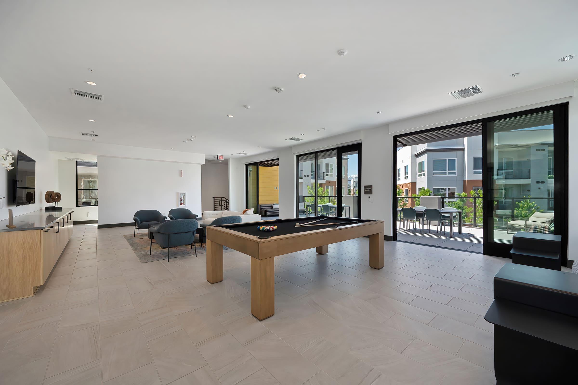 A modern lounge area featuring a billiards table, comfortable seating, and large glass doors leading to an outdoor patio. The interior has a sleek design with neutral tones and ample natural light, creating a welcoming atmosphere for relaxation and socializing.