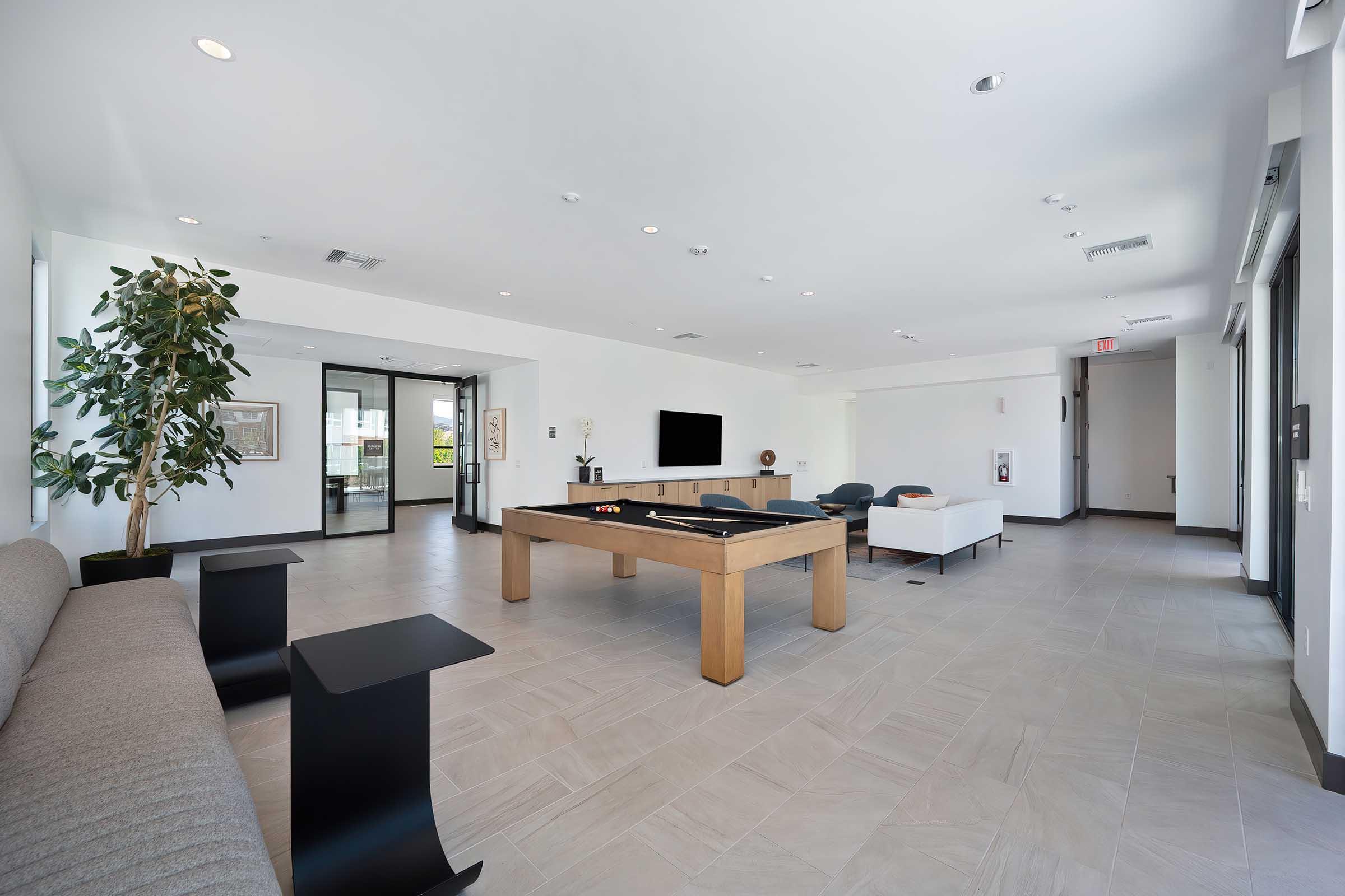 A modern, spacious lounge area featuring a pool table, a gray sofa, and a white armchair. Large windows allow natural light to enter, with a decorative plant in the corner. A flat-screen TV is mounted on one wall, and the floor is made of light-colored tiles, creating a sleek and inviting atmosphere.