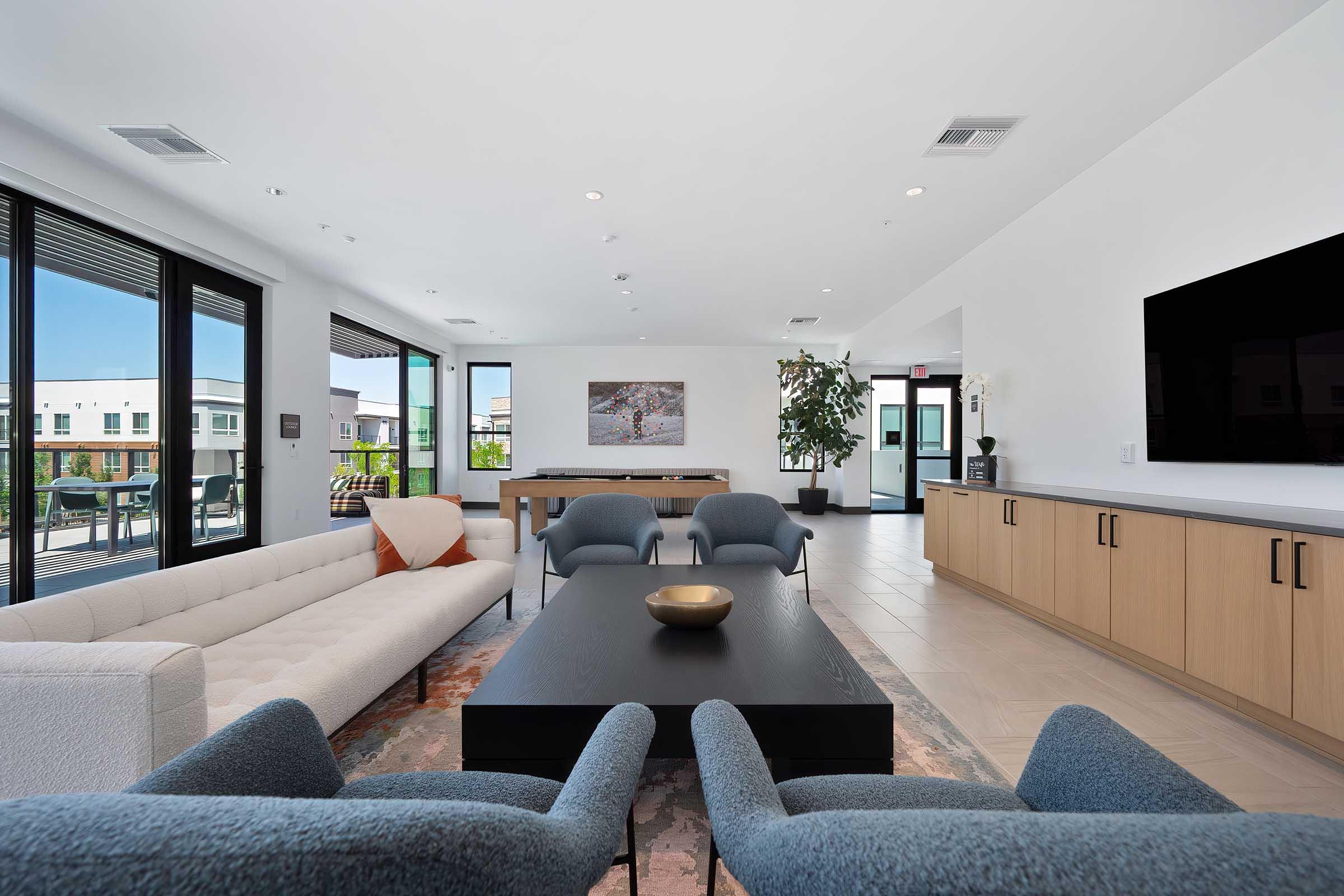 A modern lounge area featuring a light-colored sectional sofa, two blue accent chairs, and a dark coffee table. Large windows provide natural light, and a wall-mounted TV is visible. The space includes a wooden sideboard and contemporary art on the walls, along with greenery from a potted plant.